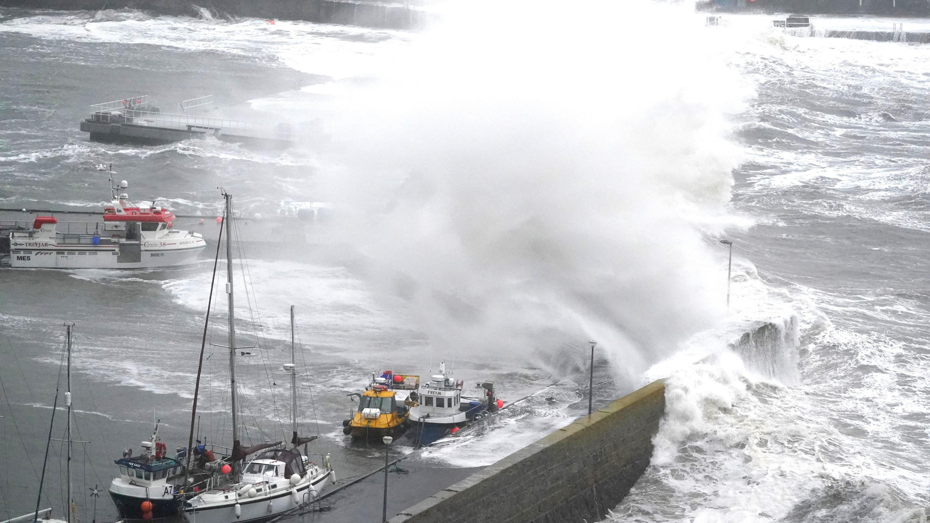 Large white waves crashing into a harbour over small boats that are tied up