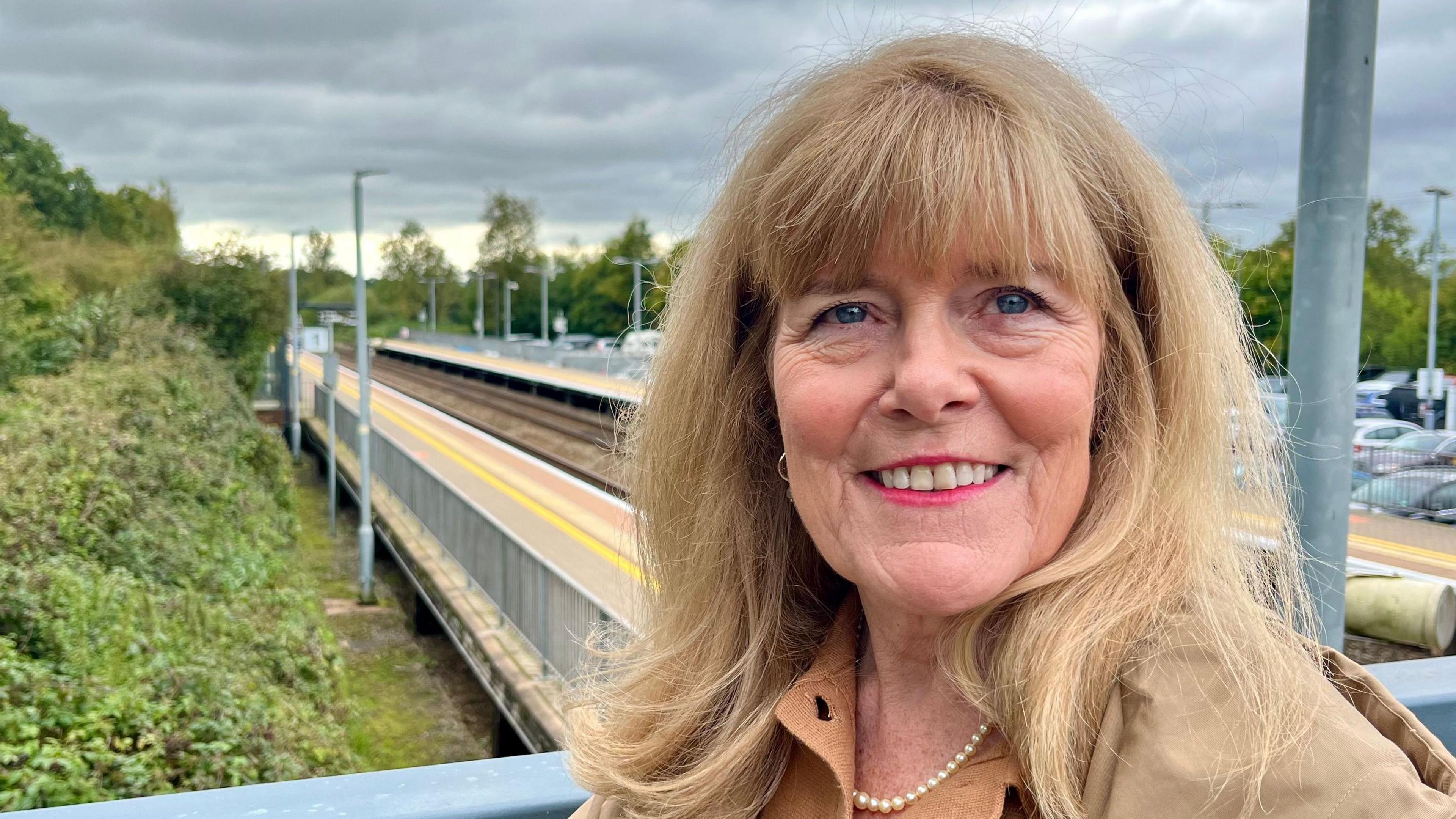 Andrea Davis is wearing a fawn top and a pearl necklace and standing in front of the platform at Tiverton Parkway