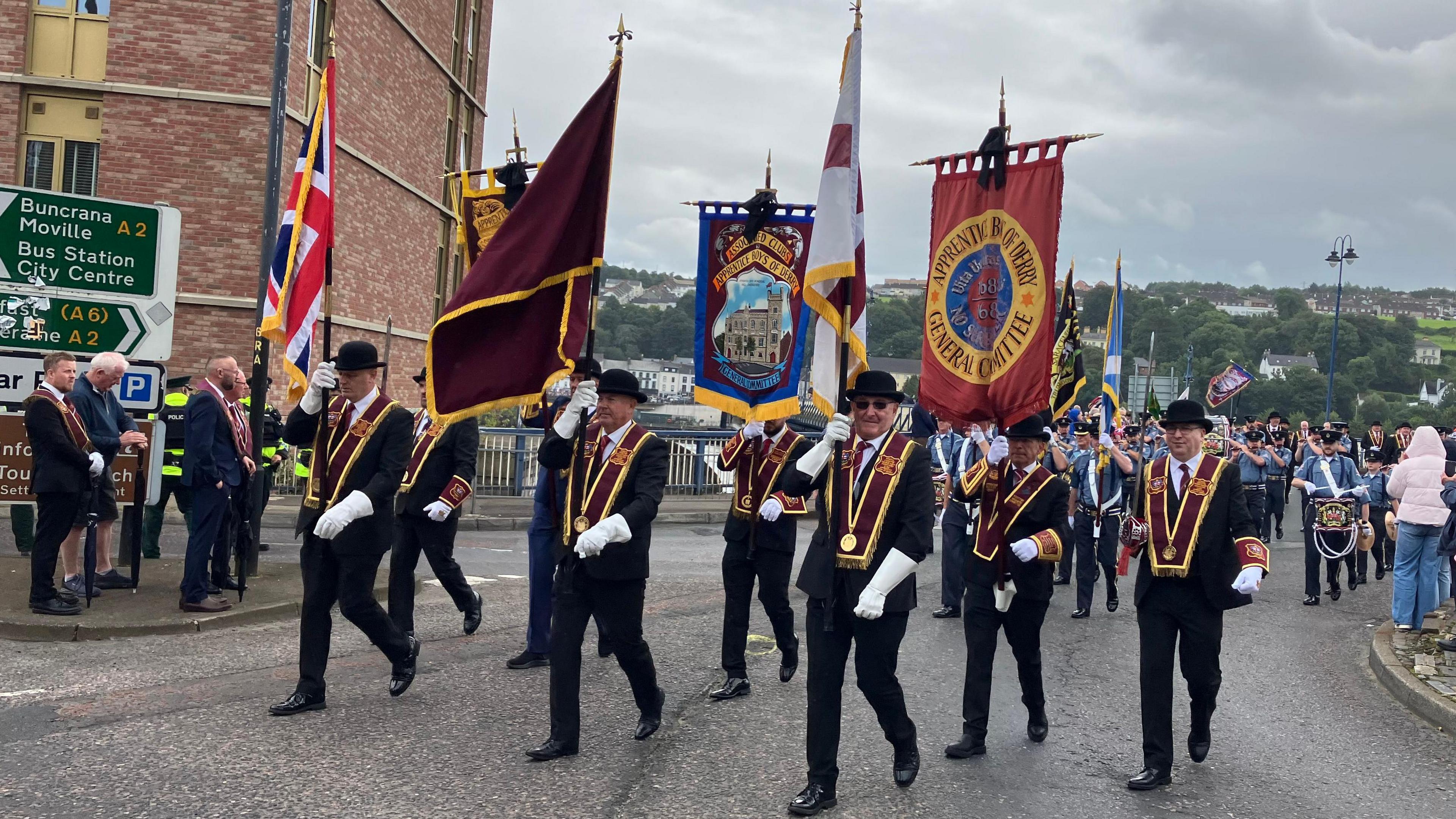 A group of men wearing black suits and black bowler hats marching in a parade. They are wearing maroon sashes an carrying various flags. There is a band behind them.
