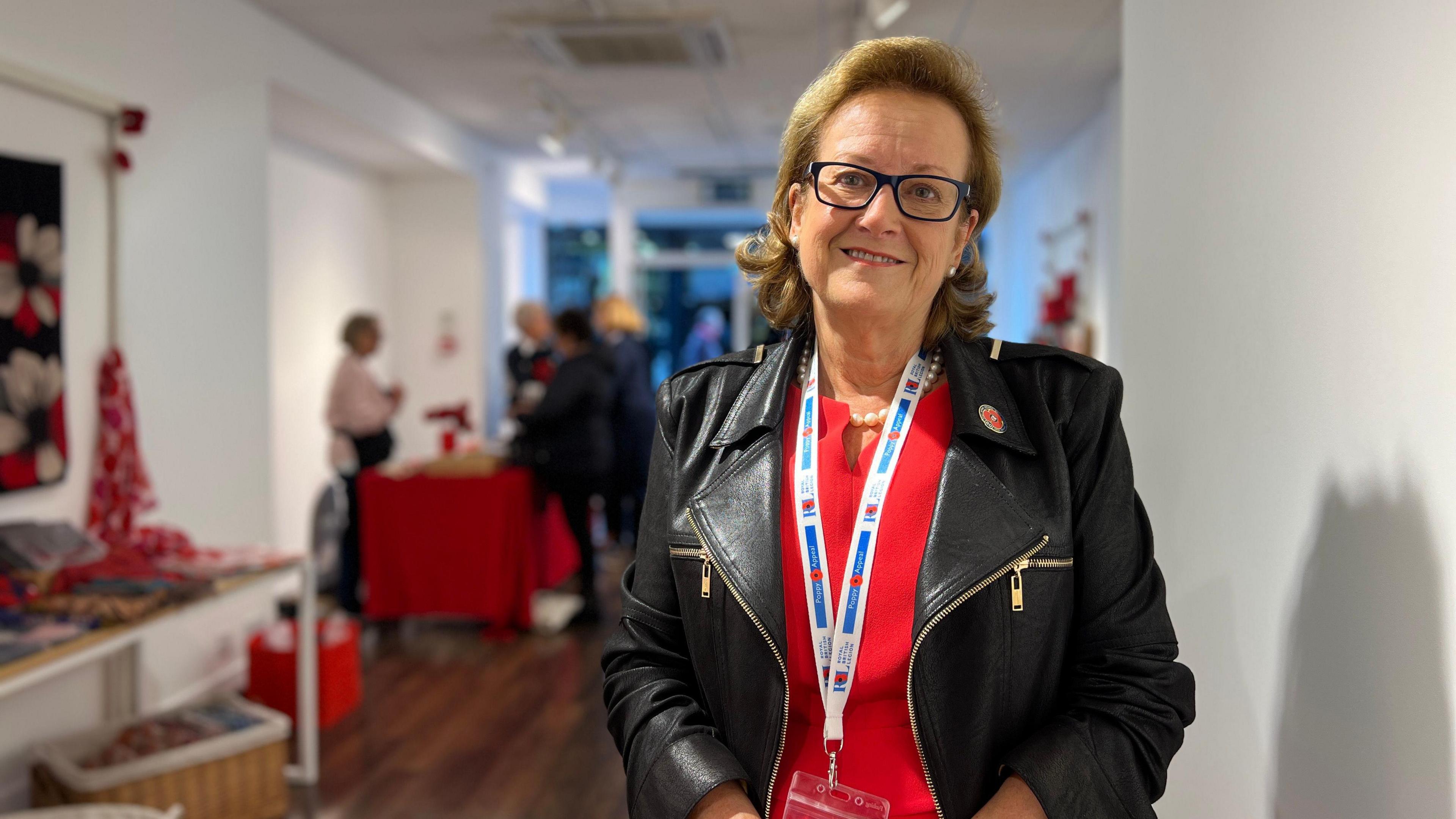 Alison smiles at the camera as she wears a red dress with a black leather jacket on and a poppy appeal pin is on the jacket. In the background people are buying poppies from volunteers in the poppy shop.