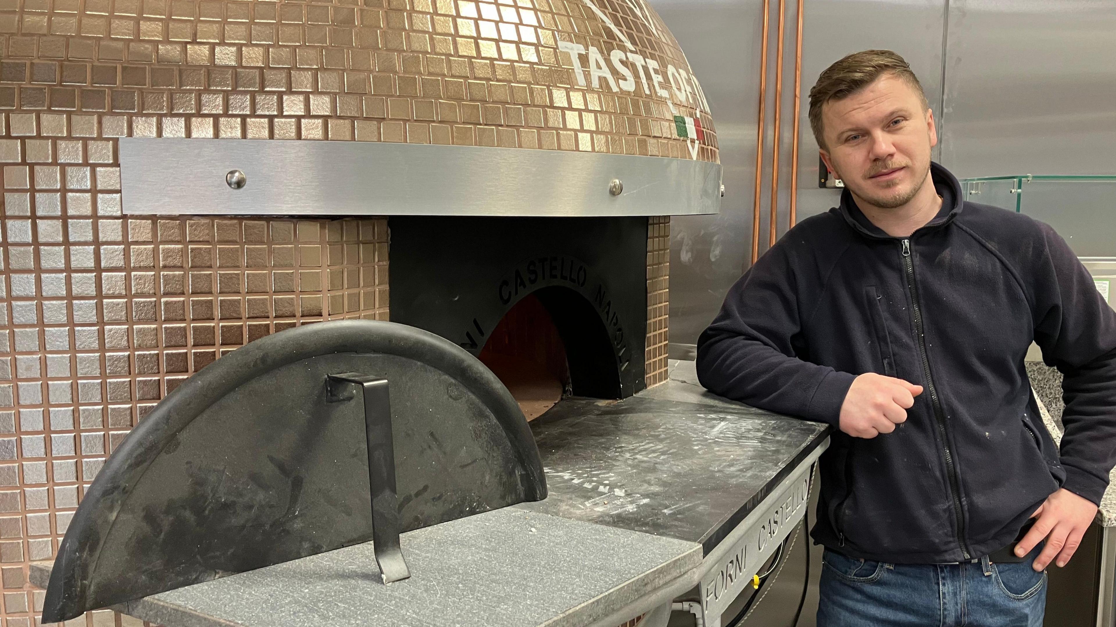 Sergio Teacu, who has short brown hair and stubble, leans on a new pizza oven. He is wearing a blue zip-up top and blue jeans.