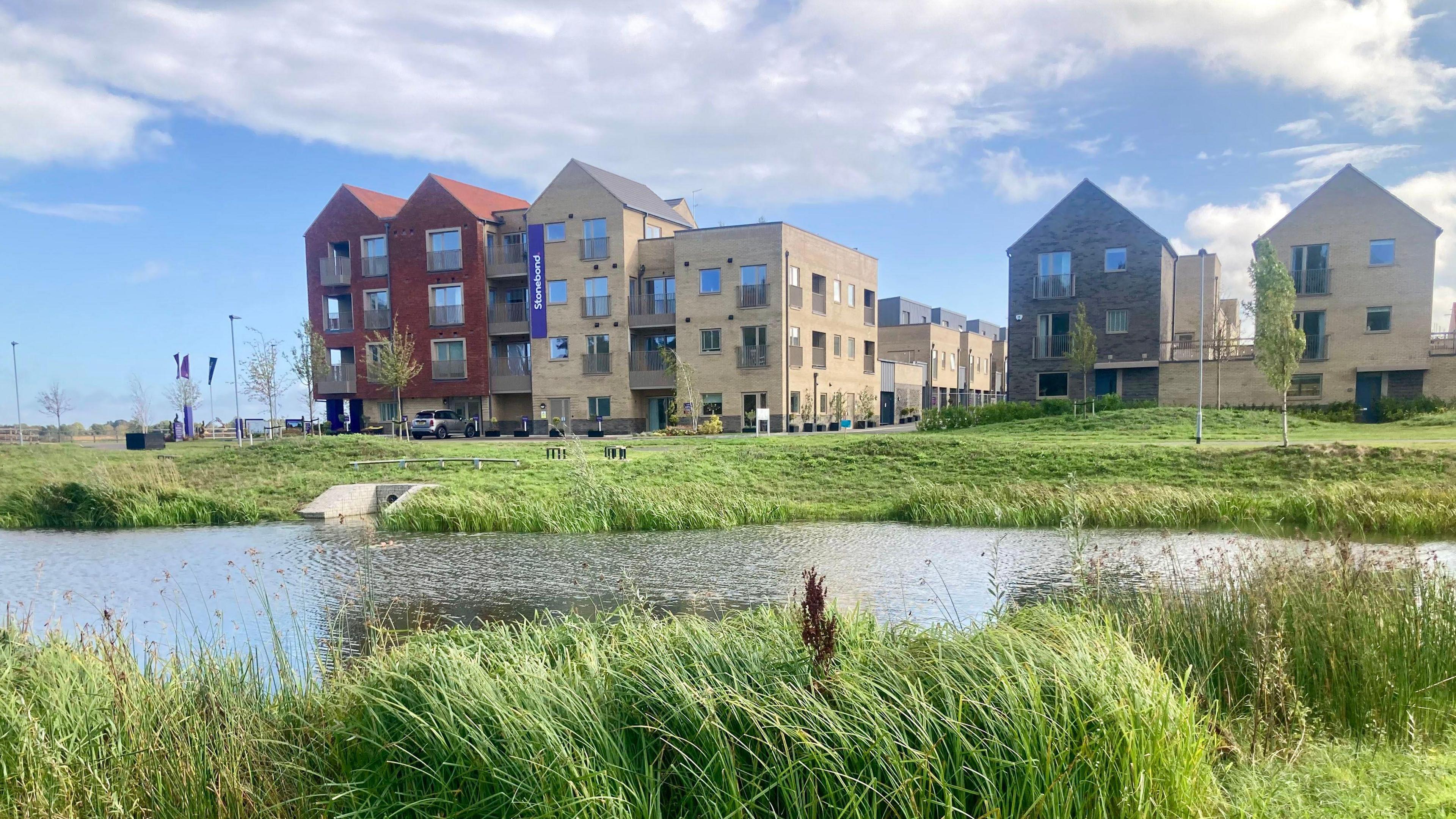 Three-storey new-builds are in the background, set against a sky that is blue and cloudy. In front is an area of grass and in the foreground is a pond with water in - an example of a sustainable drainage system.