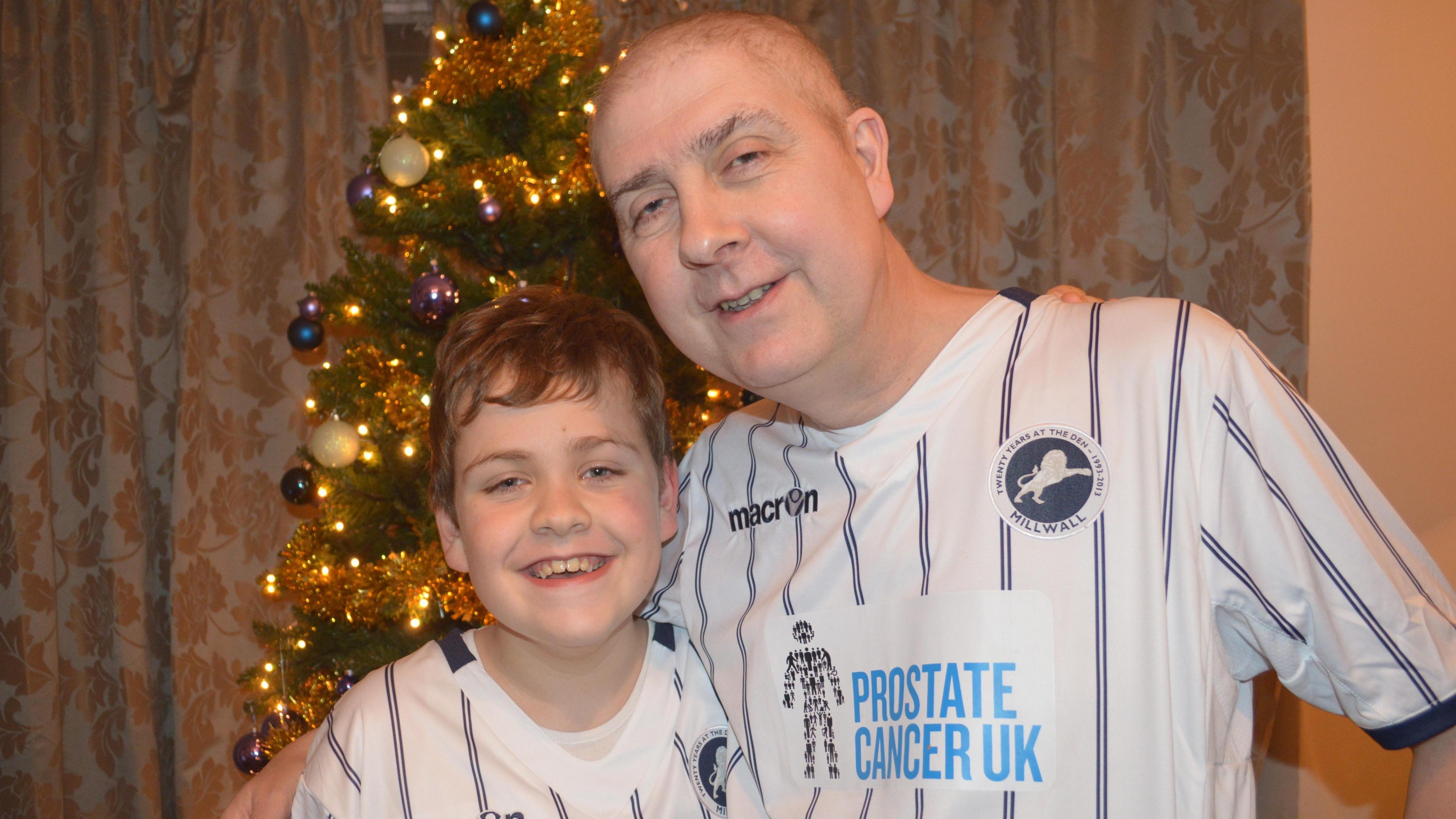 Will and Brian Kilgannon wearing Millwall Football Club T-shirts in front of a lit Christmas tree. Will is a young boy.