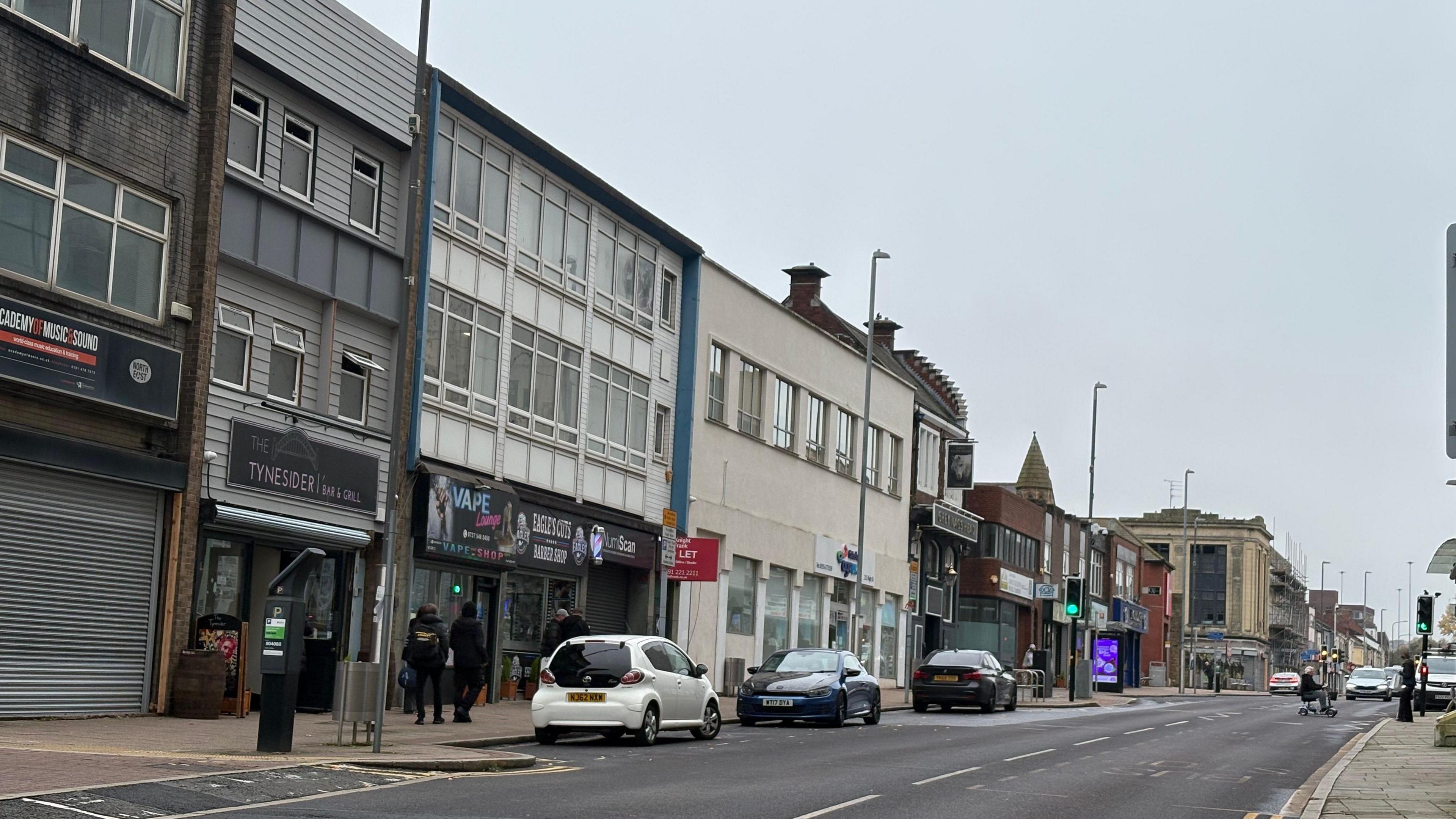 Gateshead High Street. A number of cars are parked along the street. There are pubs and shops, some with metal shutters rolled down. It looks almost empty.