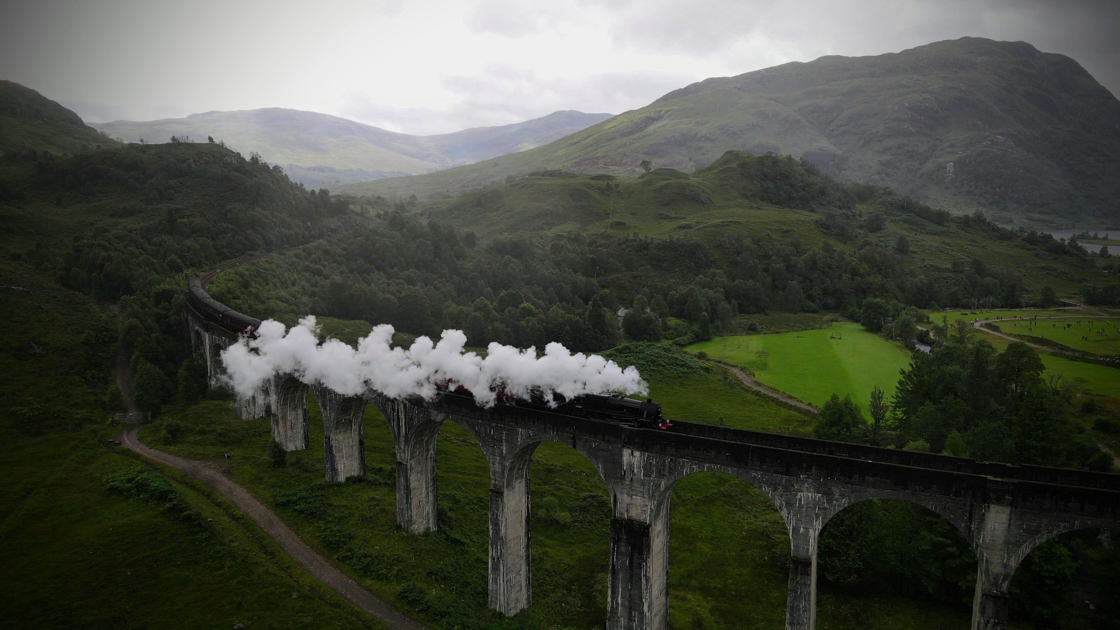 White plumes of smoke rise from a train on a train track which is elevated by stone pillars. There are green fields and mountains all around it.