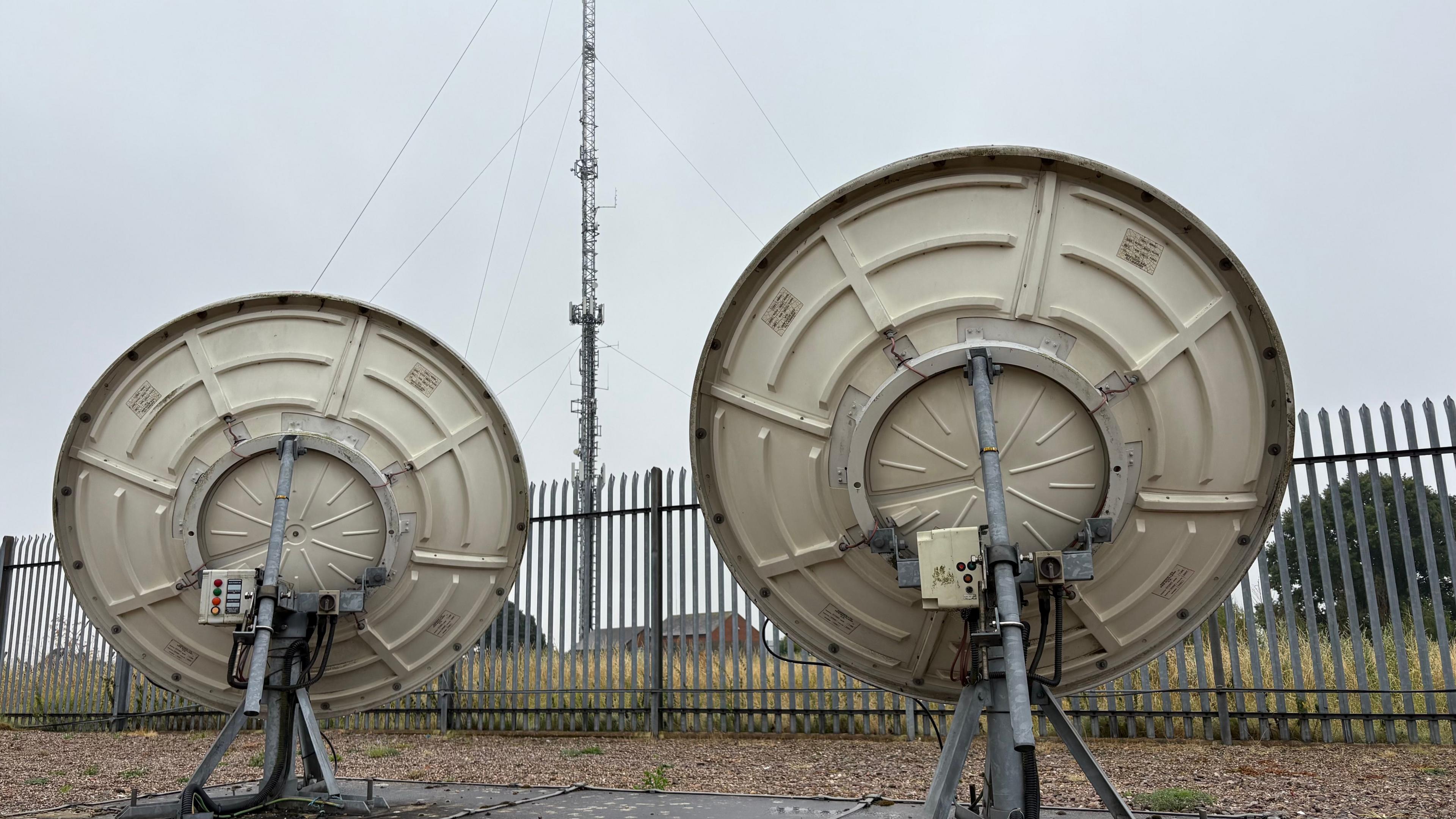 The rear of two large grey satellite dishes on metal stands. Between them, on the other side of metal railings, is the lower section of a mast, on which small aerials can be seen. There are wires tethering the mast to the grass below. There are buildings and trees in the distance. 