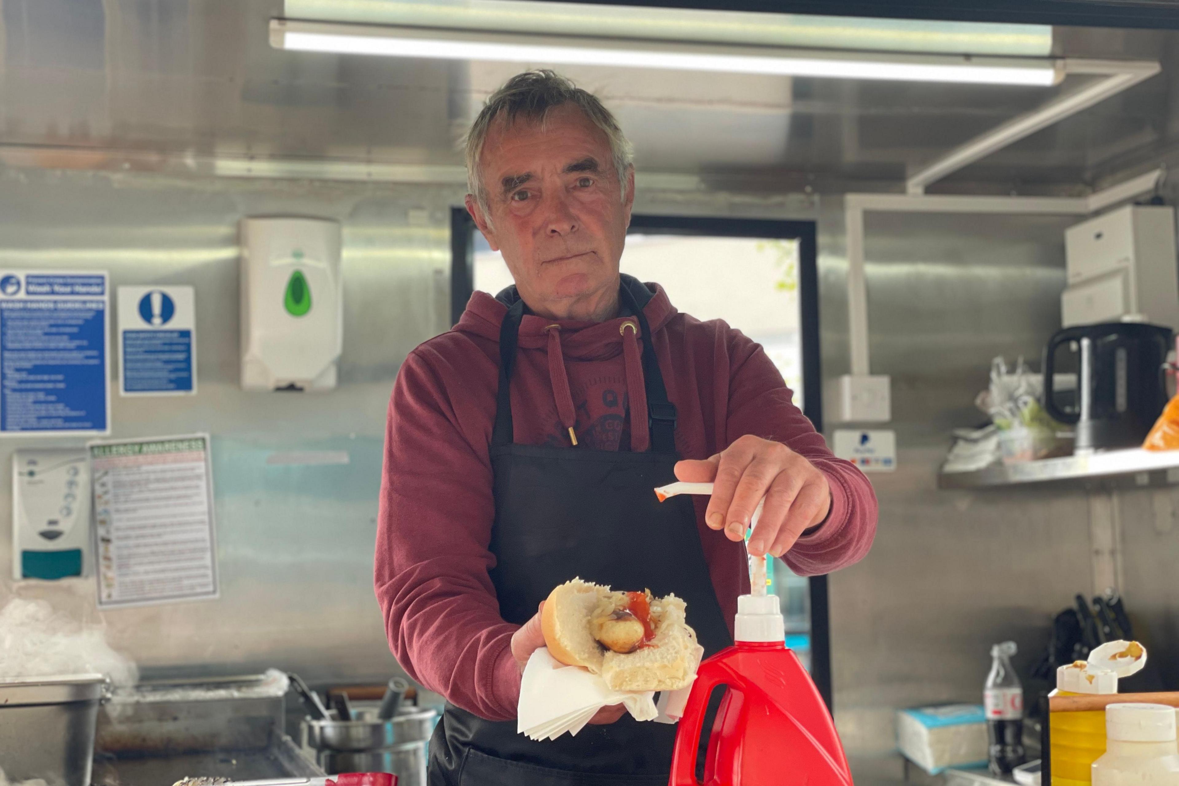 Charlie Pearson looks into the camera as he stands in a kitchen area, wearing an apron and holding a long bread roll with a hotdog inside it. His other hand is resting on the top of a large red ketchup bottle with a white dispenser pump. 