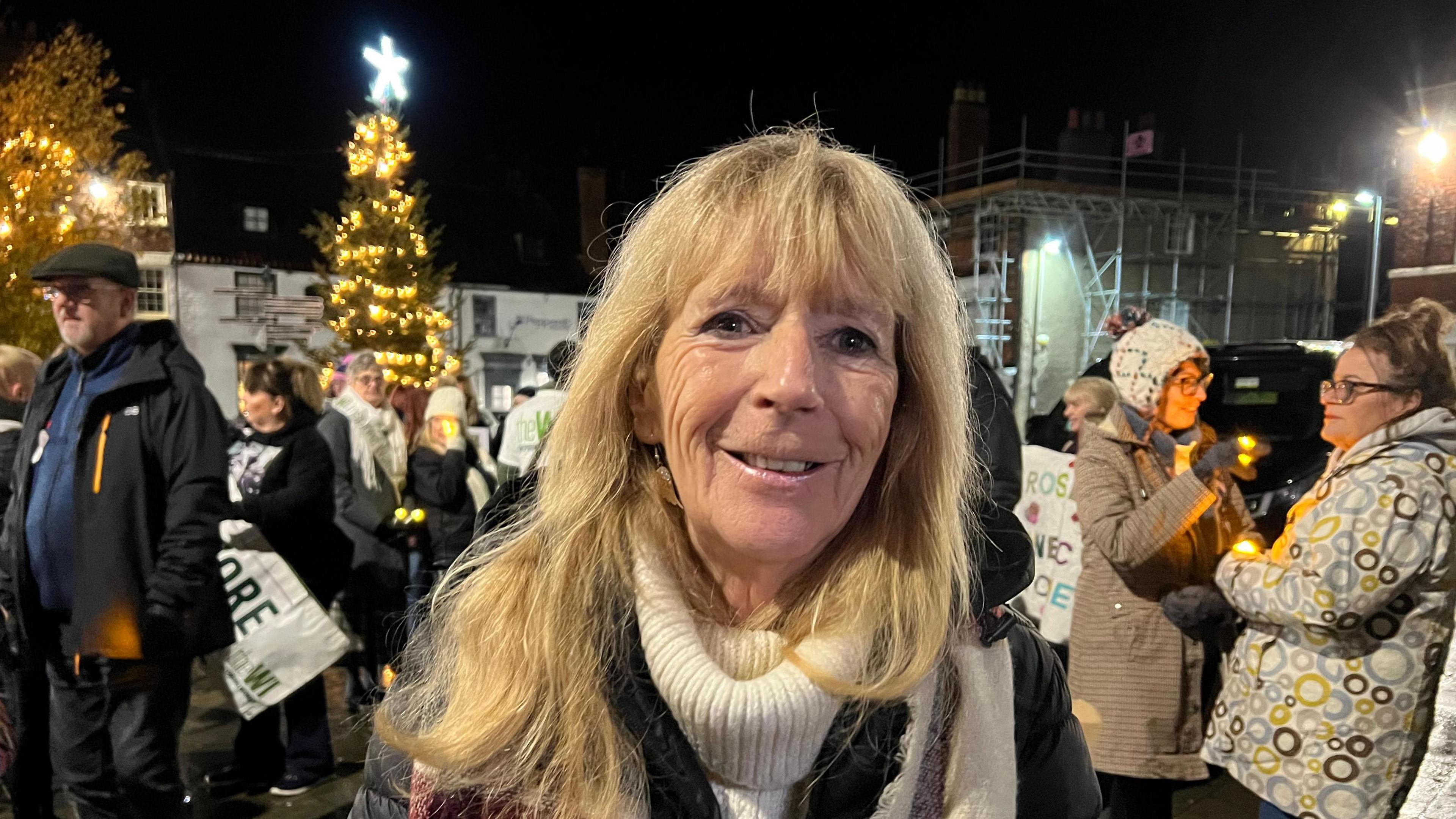 Woman with blonde hair and white jumper wearing a dark coat. There is a crowd behind her.