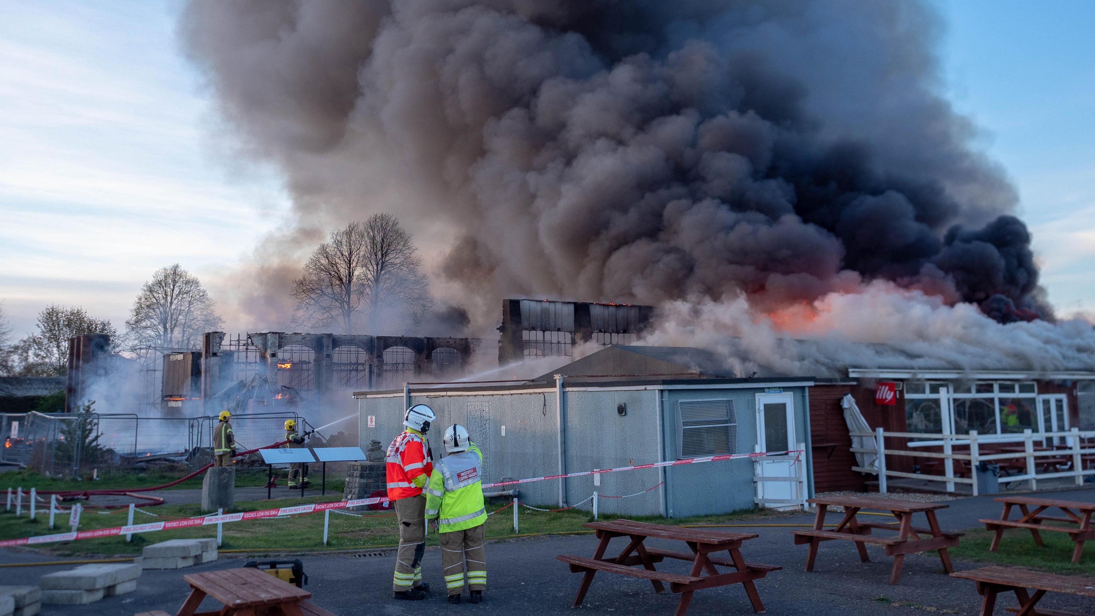 Two firefighters aim water at a huge building on fire, while two others wearing high vis clothing look on. The building is clearly engulfed by fire, and there is a huge black plume of smoke rising from it, as well as flames. 