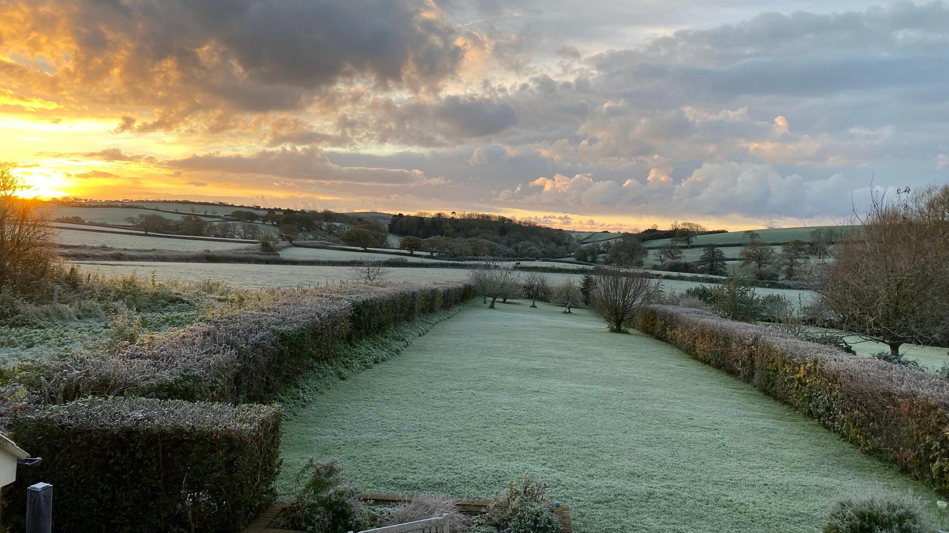 Frosty looking fields and hills with a low orange sun shining on the glistening grass below.