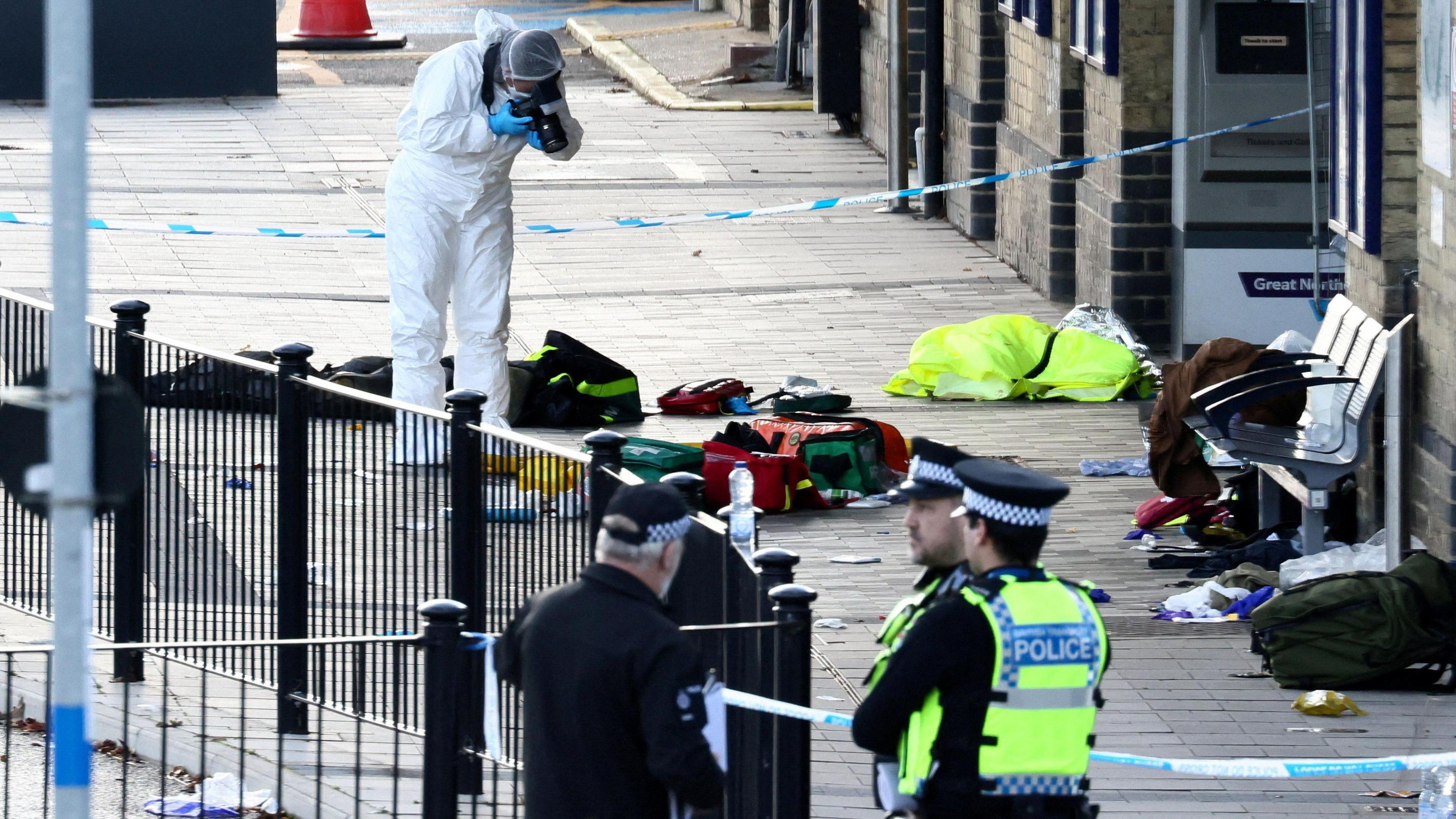 An area of Huntingdon railway station cordoned off. There are police officer standing outside the cordon. However, a forensics officer stands within the cordon photographing items inside of it.