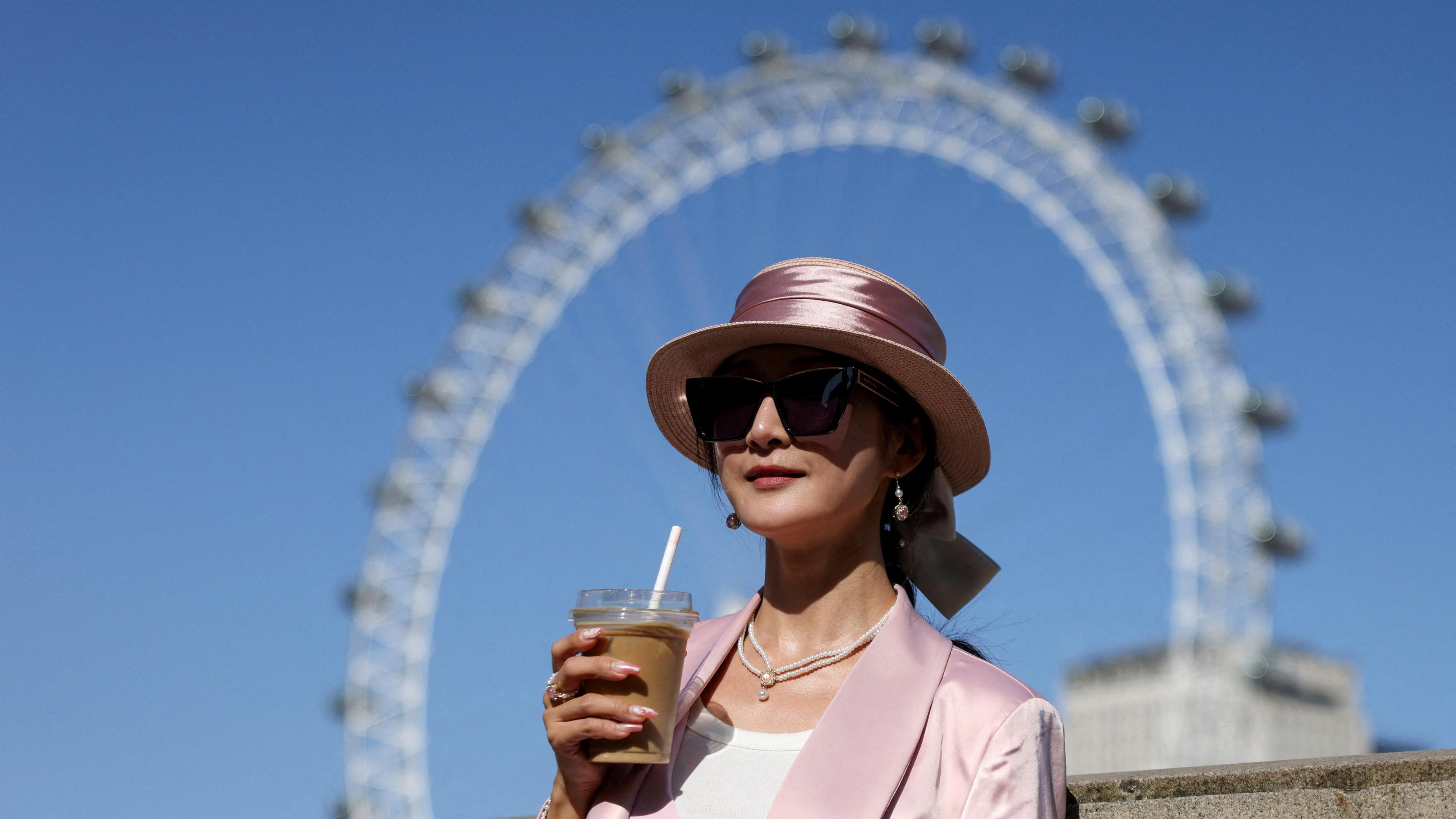 A woman holds a drink while posing for a photo in front of the London Eye, in London