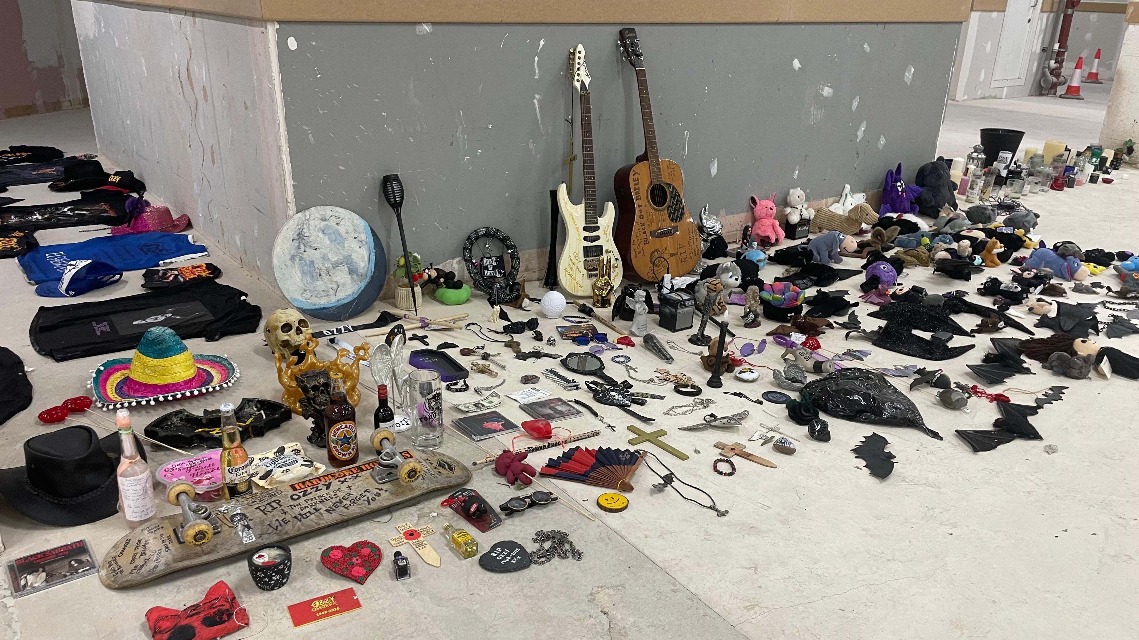 Dozens of objects laid out on the concrete floor of a warehouse. They include two guitars, dozens of cuddly toys, and several football shirts. There is also a skateboard, a skull, and a colourful sombrero.