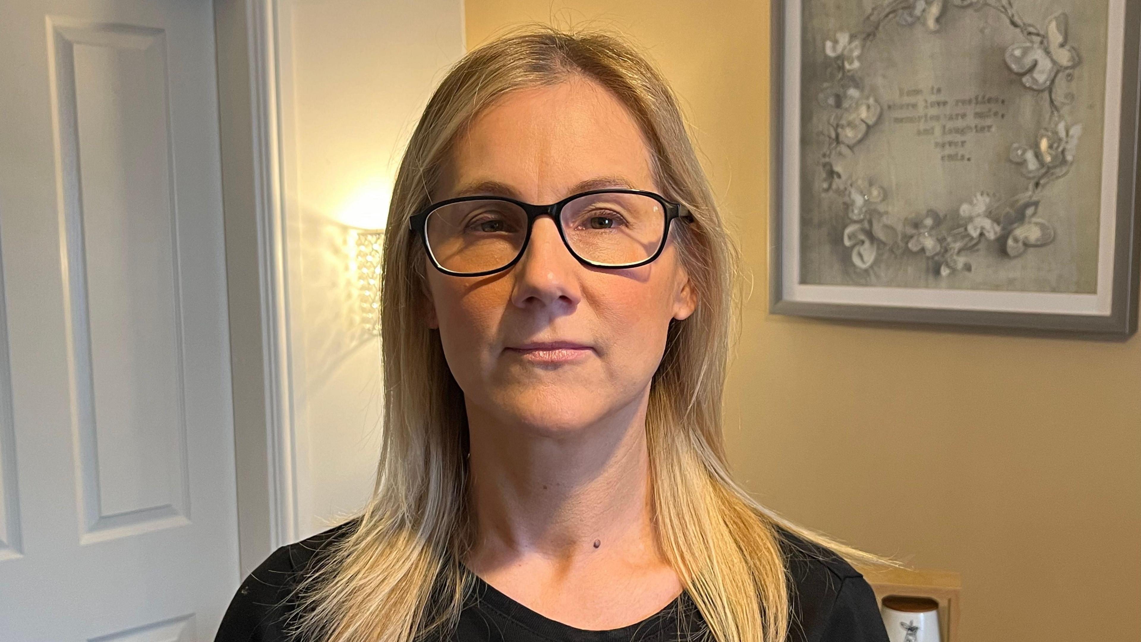 Head and shoulders of a woman with blonde hair in dark framed glasses and a black top. She is standing in her home, with a light and sketch in a frame on the wall behind her