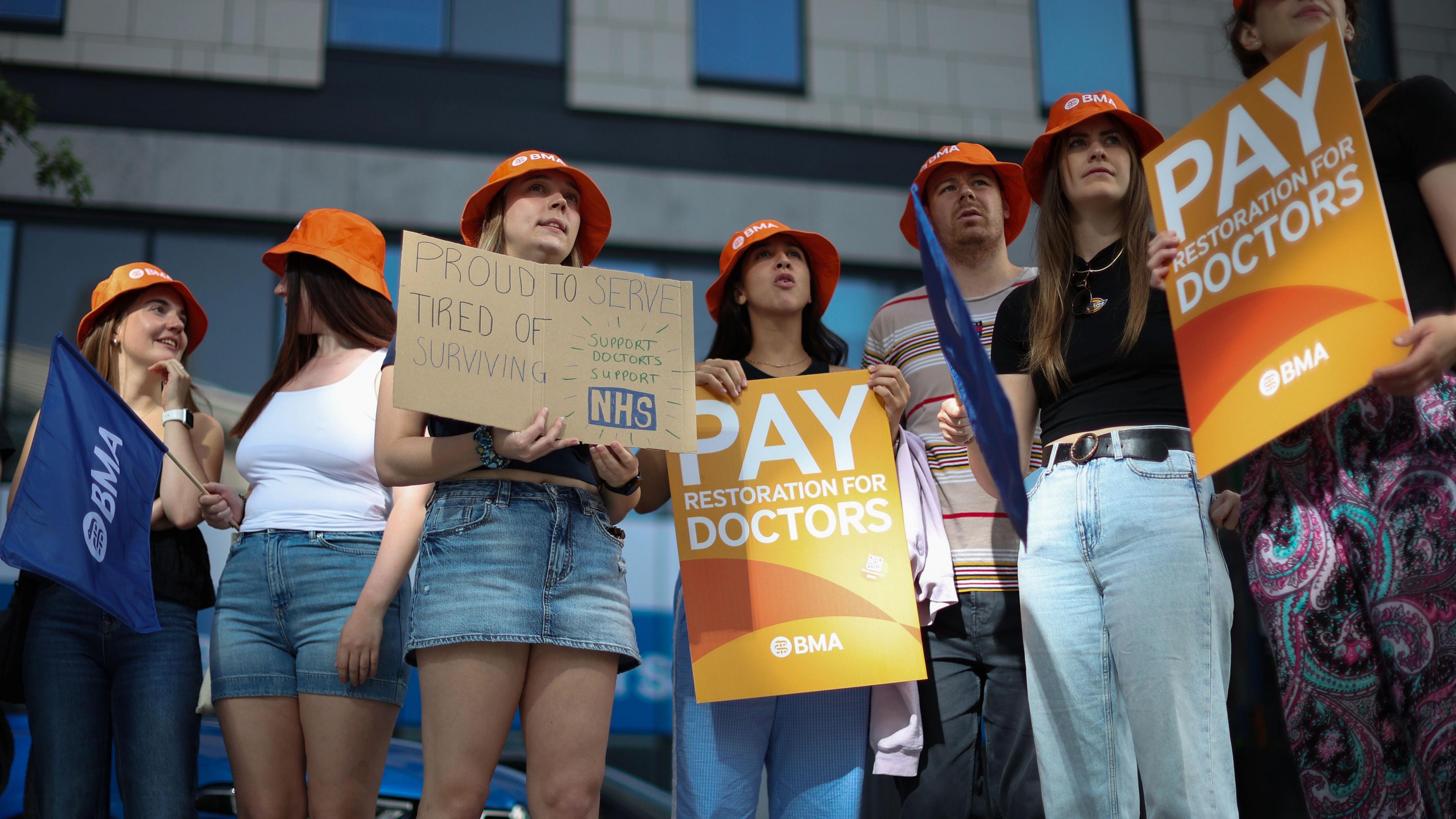 a group of doctors wearing bright orange hats and holding up placards which say proud to serve tired of surviving and pay restoration for doctors.