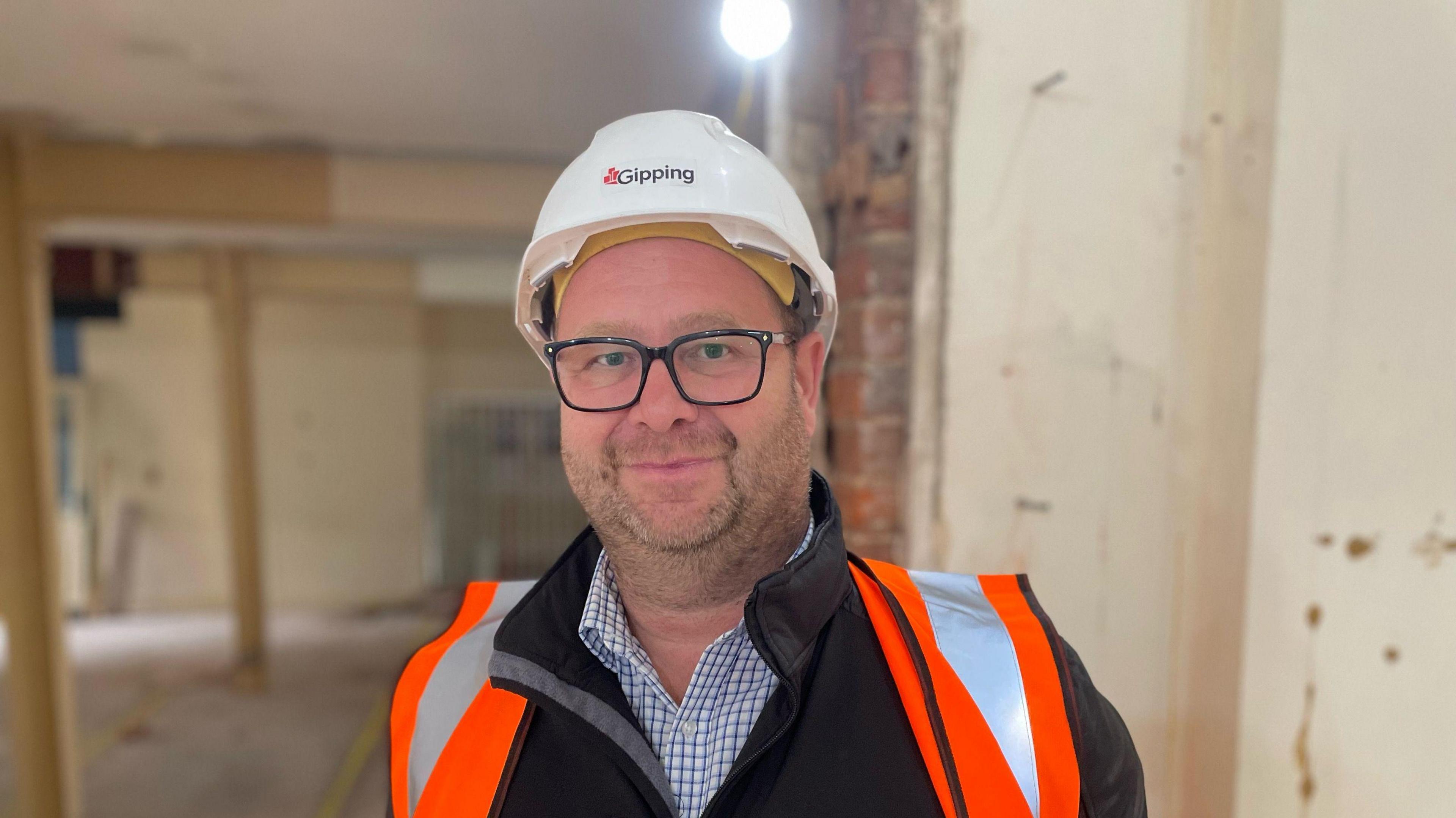 Steve Offord is pictured. He is facing the camera and smiling. He is wearing a white safety helmet and orange high-vis jacket on top of his black jacket and blue and white chequered shirt. He has black glasses and some stubble on his face. Behind him is a room in the museum which is blurred.