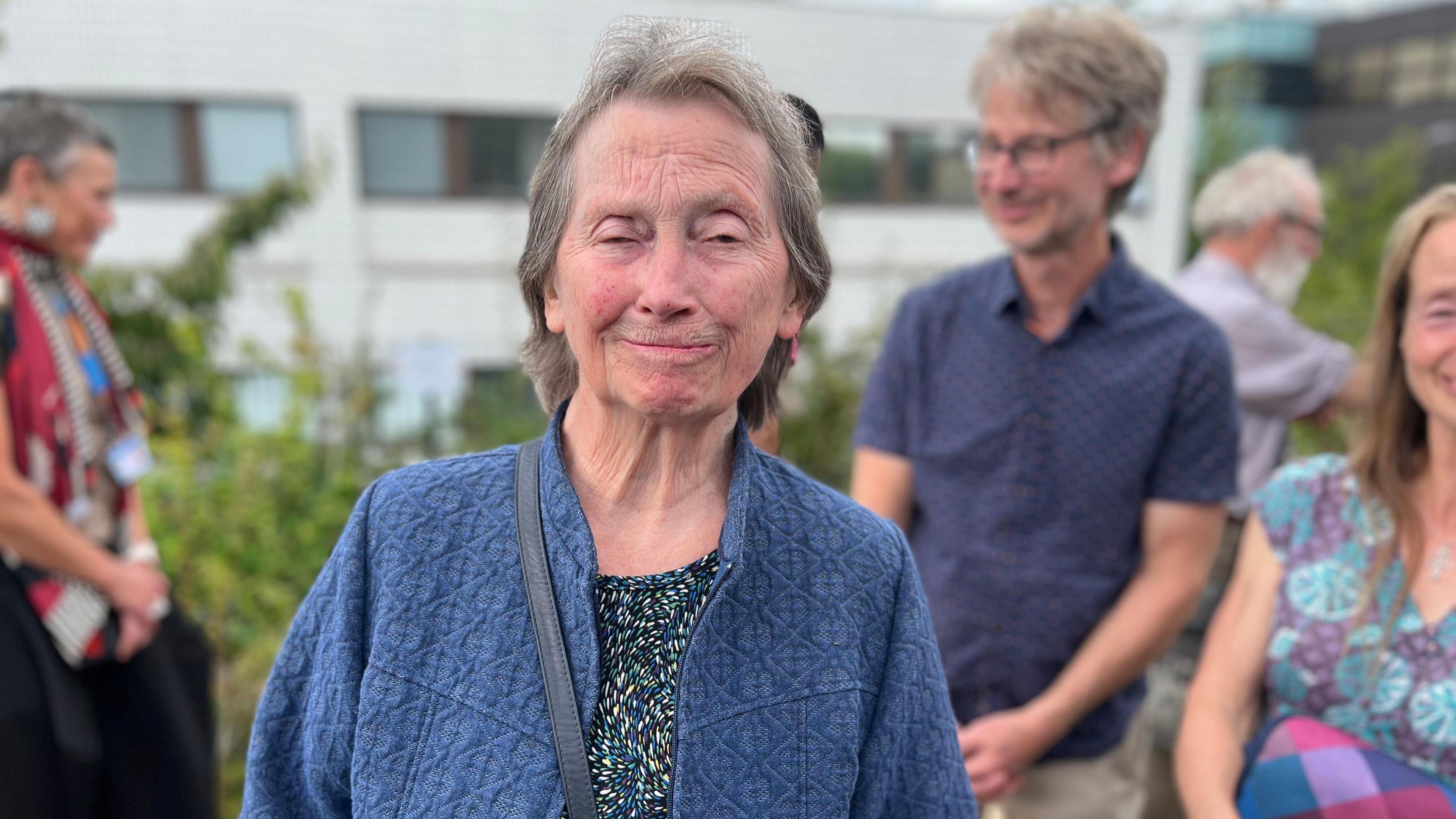 A woman with grey hair and a blue jumper smiles at the camera