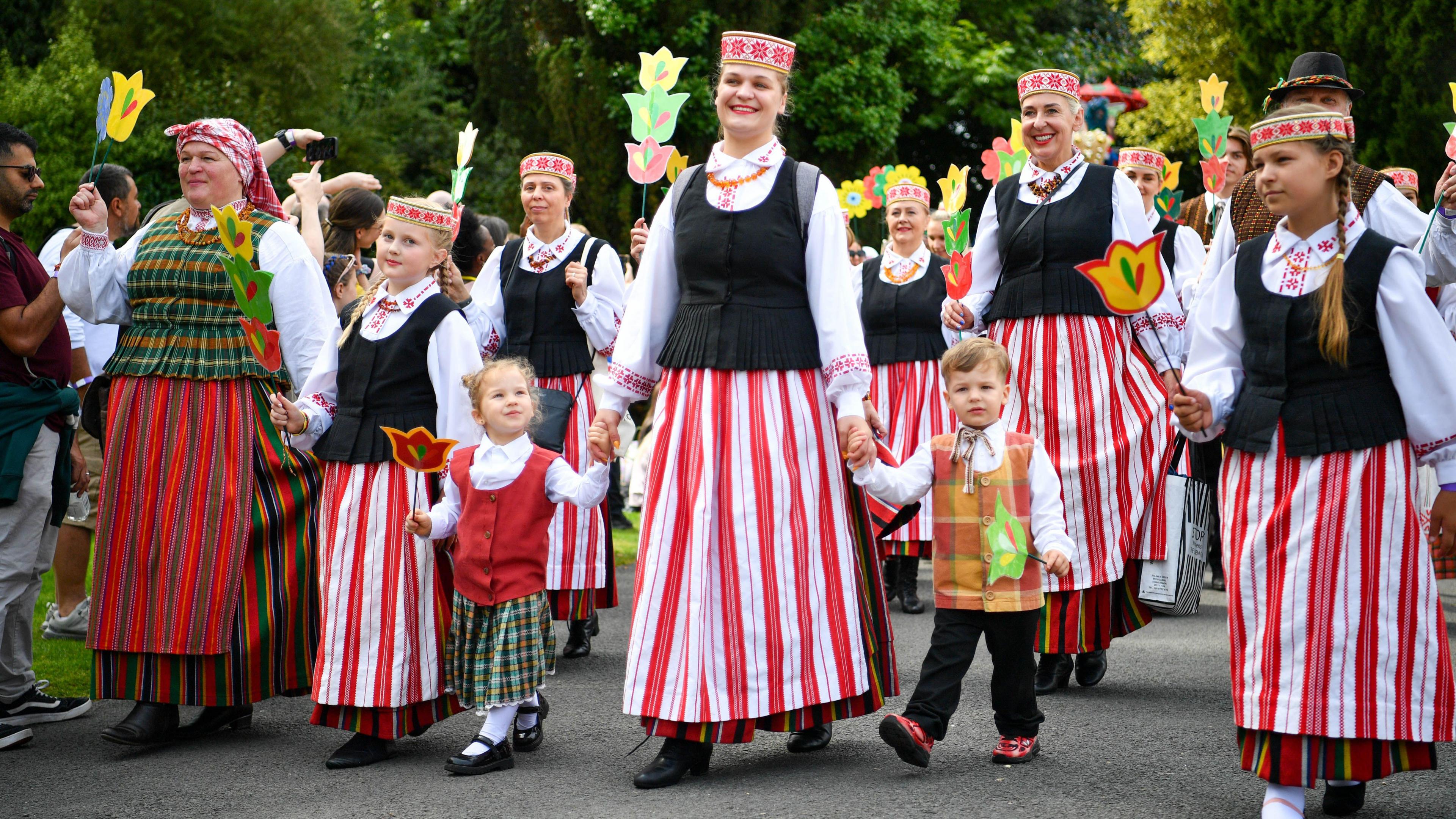 People are dressed in traditional dress and walking in a parade through Botanic Gardens. They are wearing hats decorated with red flowers. Some are holding floral decorations. 