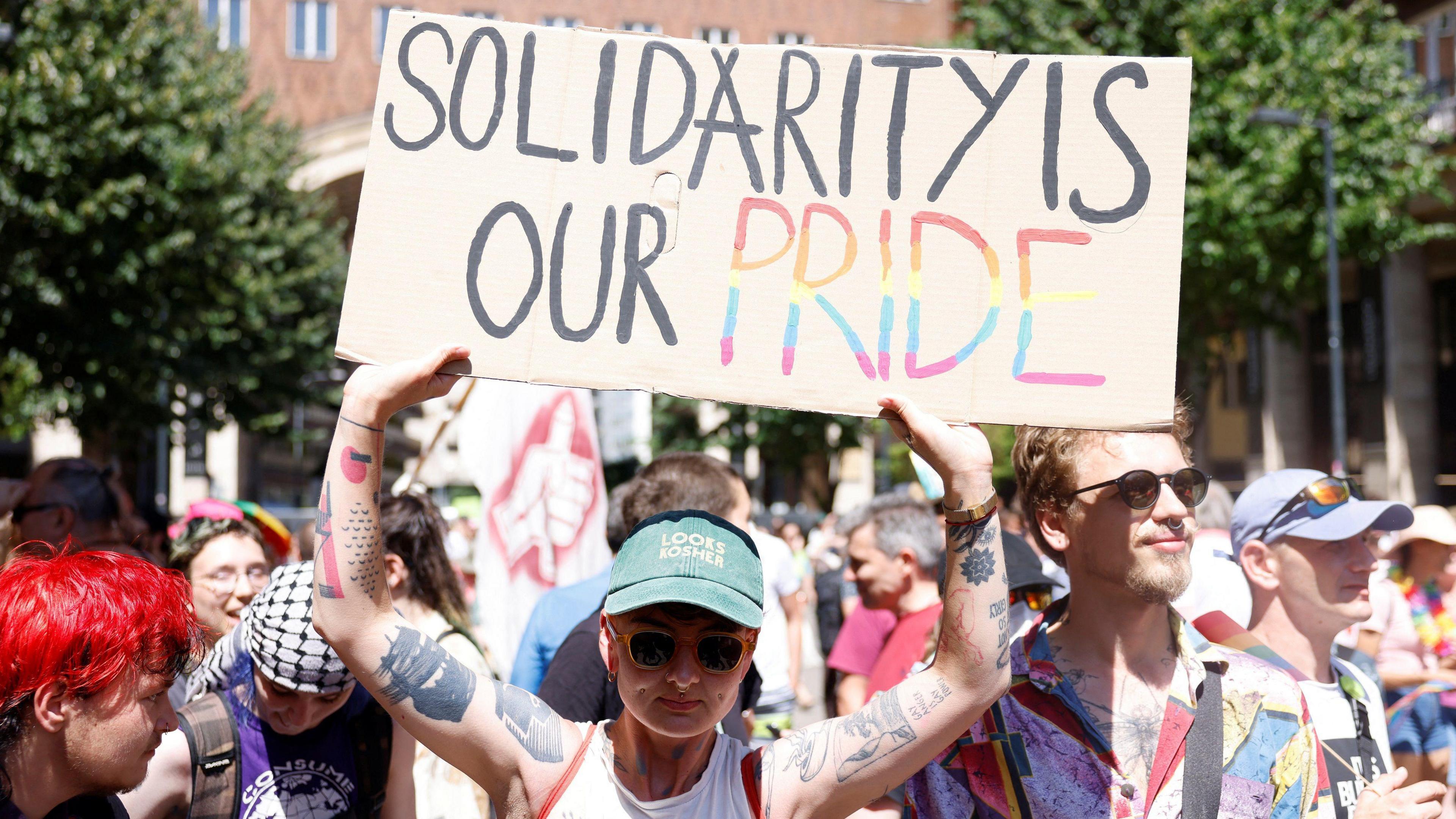 Someone standing in the march holding up a cardboard placard reading solidarity is our pride. The lettering is in black except for the word pride, which is multicoloured - red, orange, yellow, blue and purple - in horizontal lines.