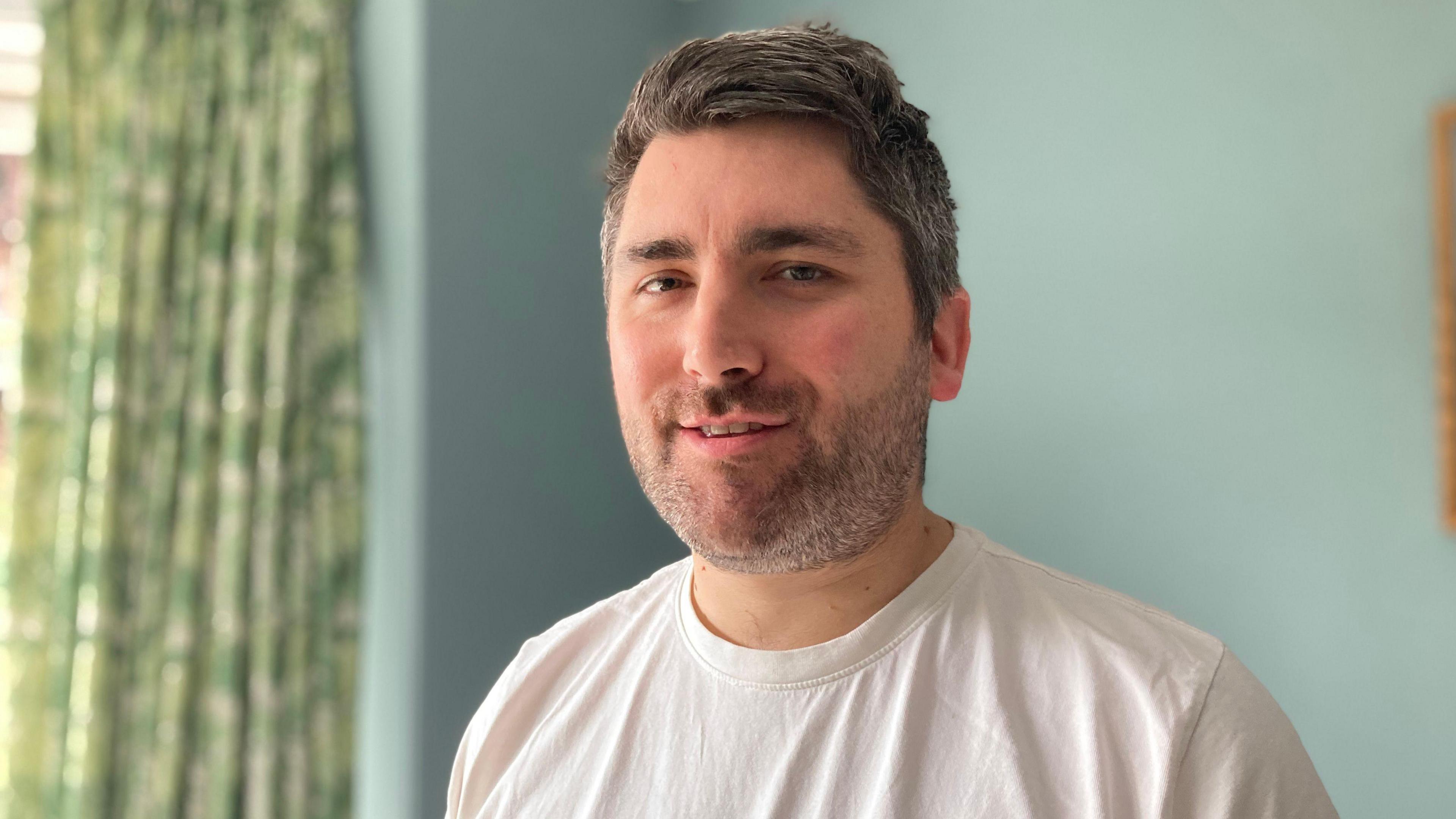 A man in a white t-shirt stands in front of a blue wall. He has facial hair and his lips are slightly parted as he smiles. His salt and pepper hair is combed over to one side.