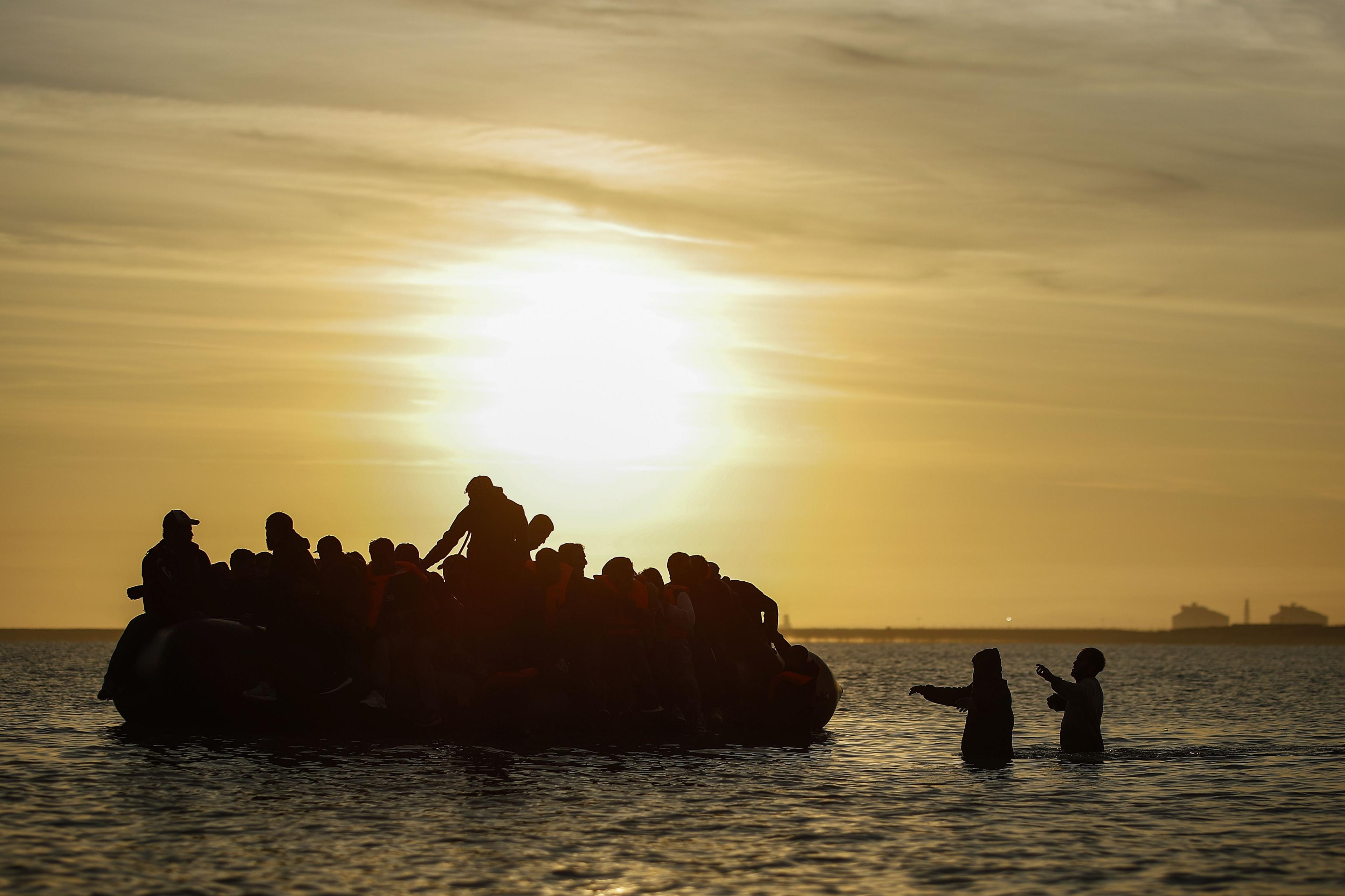 A silhouette of a dinghy carrying multiple people with two would be passengers standing in the water trying to board