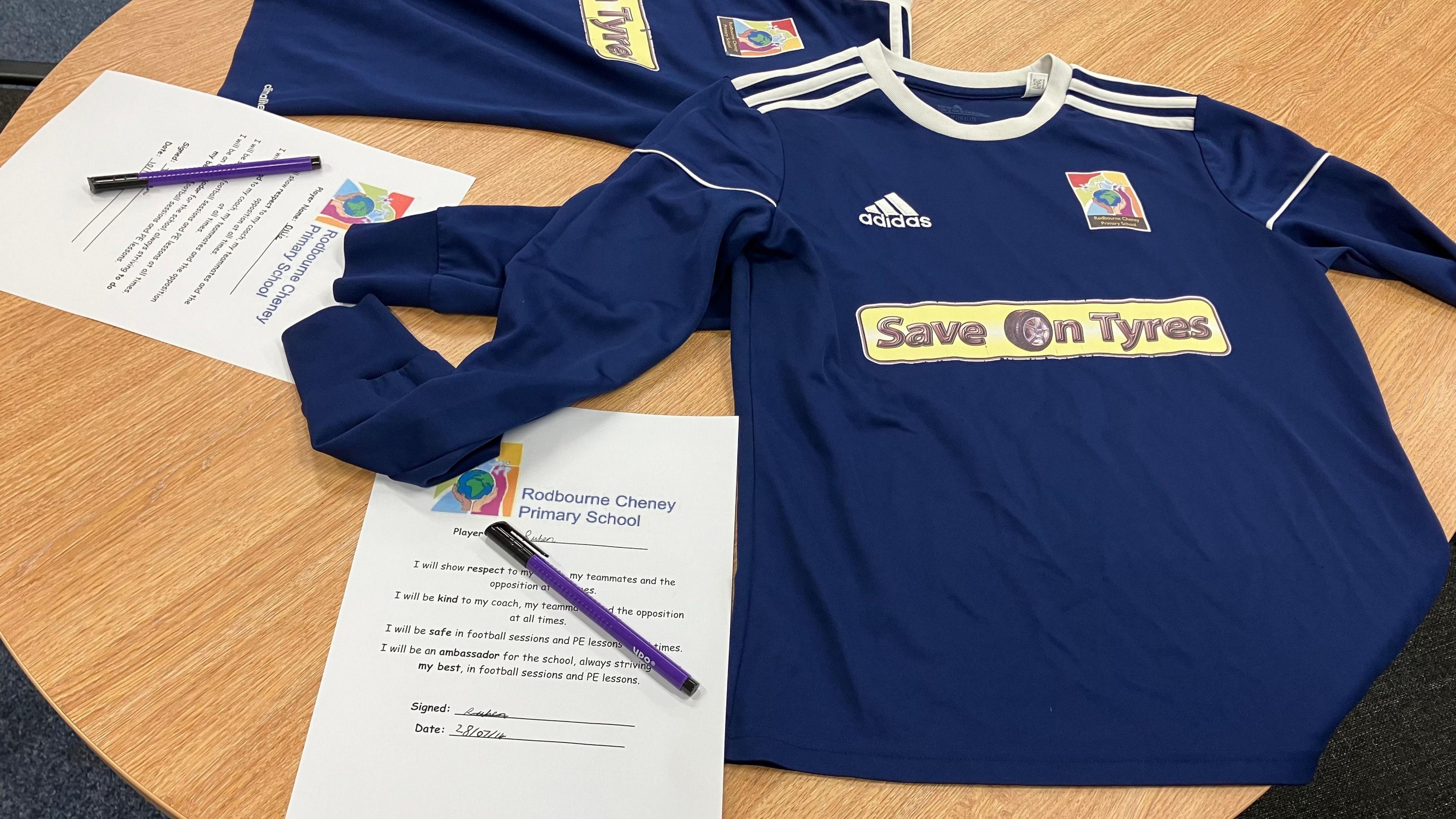 Two football shirts laid out on a table beside two football team contracts. Each contract has a pen placed on top.