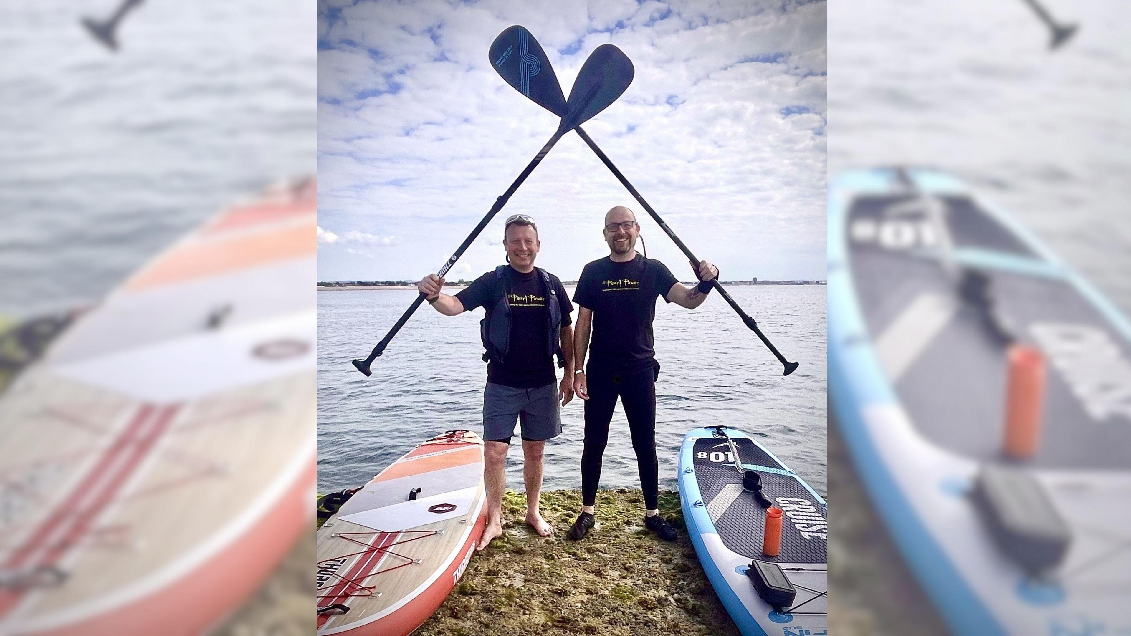 Two men holding paddles stand in front of the sea. A paddle board lies beside each of them