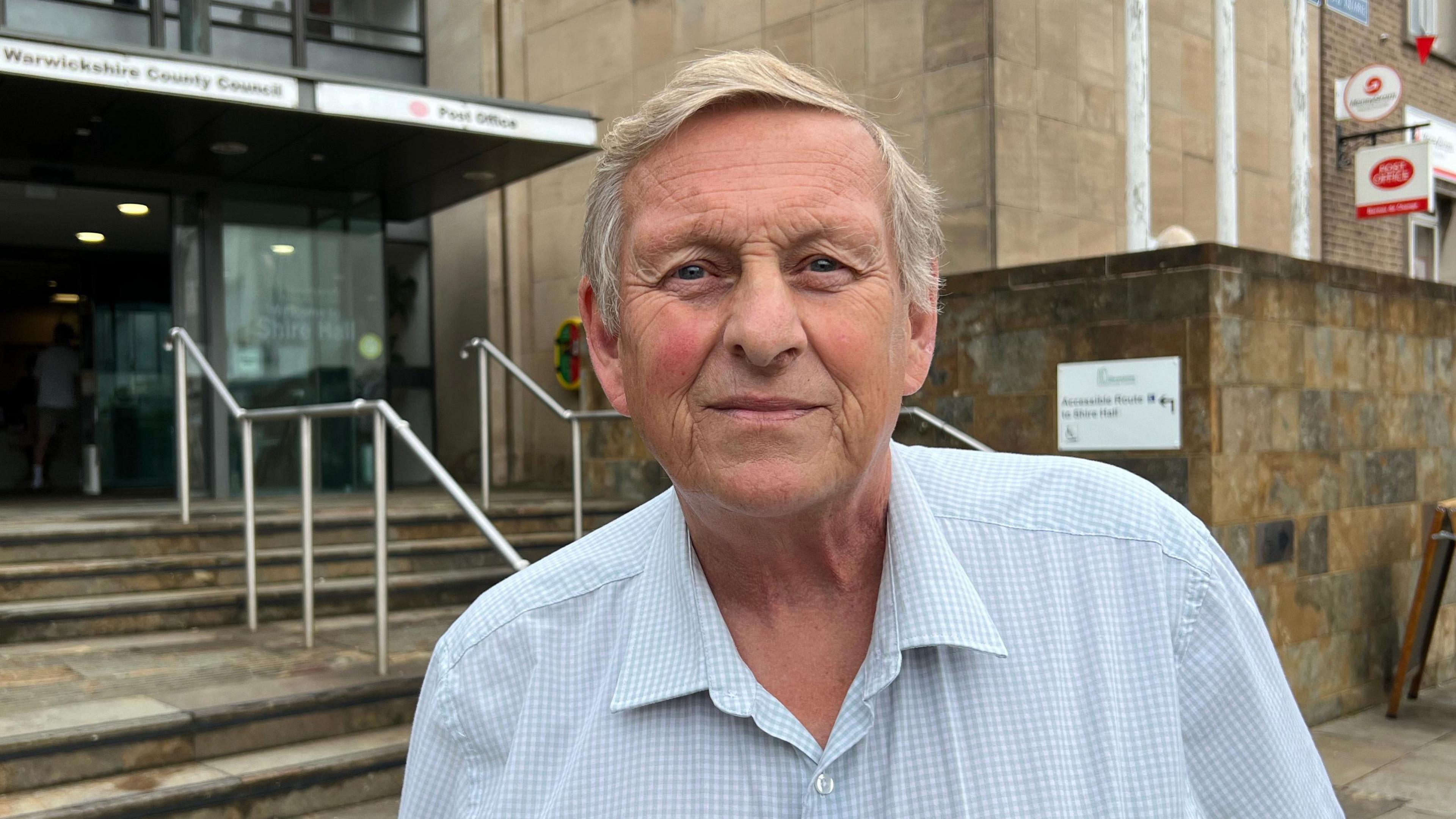 Councillor Jerry Roodhouse, leader of the Liberal Democrat opposition, stands outside the stone steps entering Shire Hall. He is wearing a blue and white chequered shirt with an open collar.