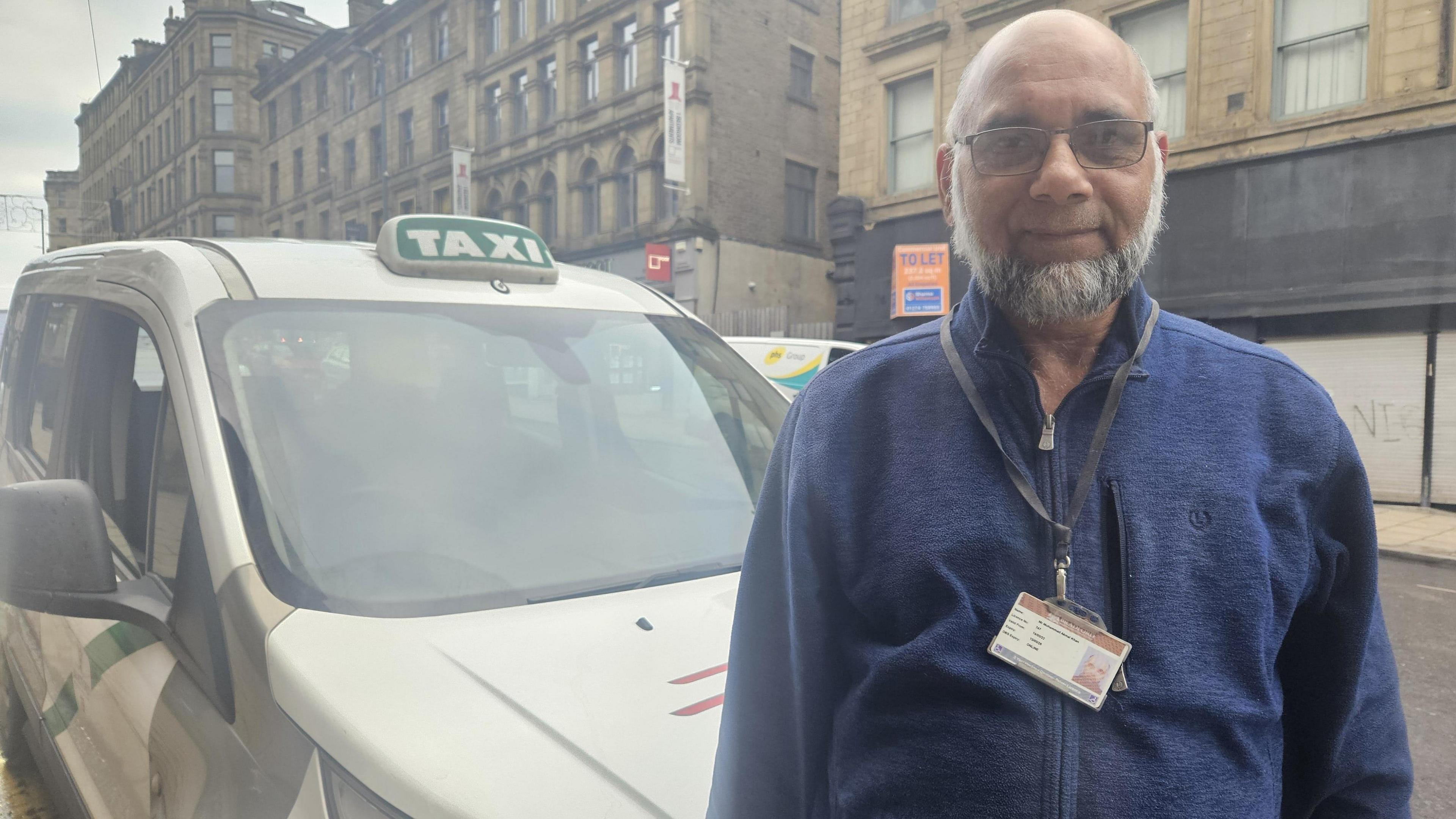 A man in a blue shirt, with a grey beard, stands in front of a white taxi, with a streetscape visible in the background.