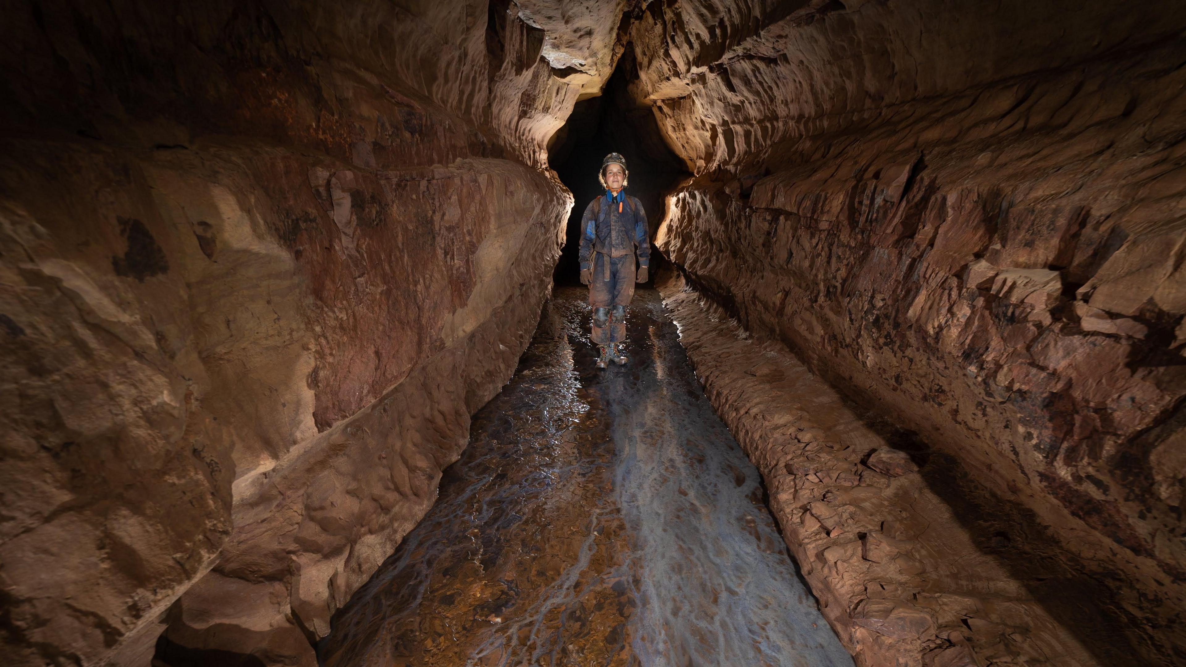 On caver wearing waterproofs and a helmet with a light on standing at the entrance of a corridor of a ricky passage. She is walking through water and looks small because of the perspective and her distance from the camera.