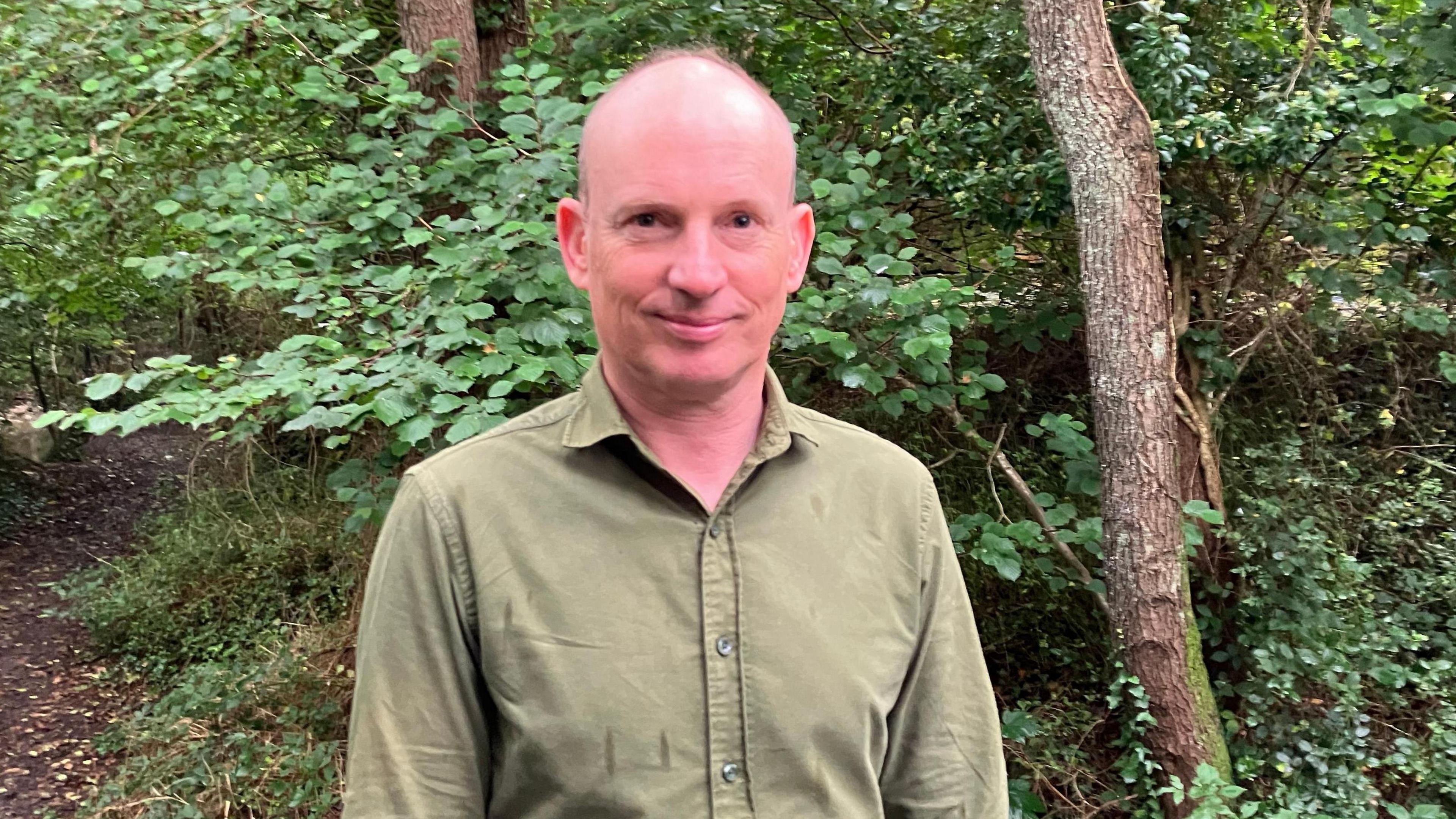 A bald man wearing an olive coloured shirt standing in front of a wooded area