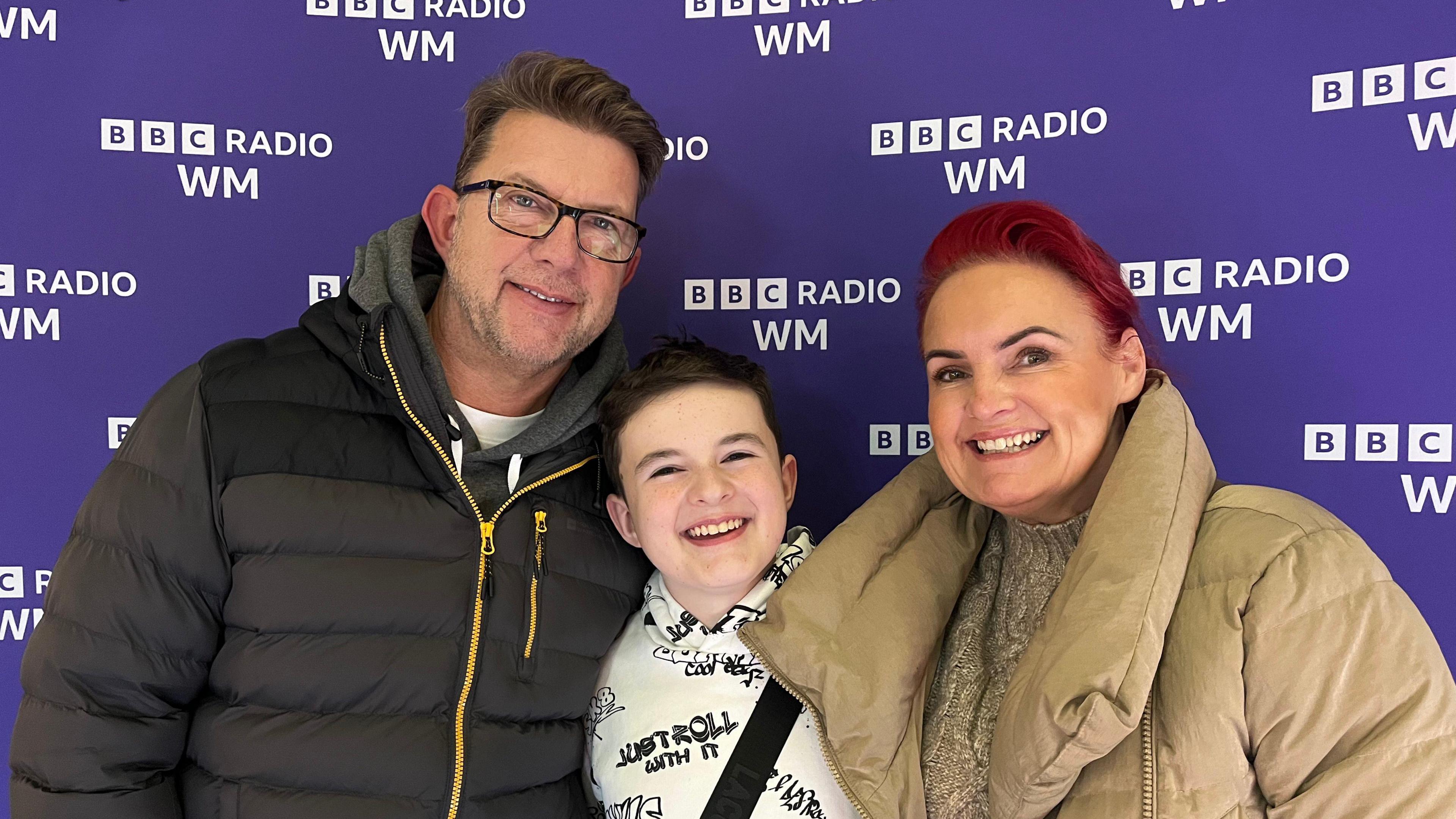 Finley Hill, with his parents, stood against a purple BBC Radio WM banner. His dad is wearing glasses and a dark grey hoodie with a yellow zip, and his mum is wearing a thickly-padded, beige-coloured coat and beige coloured jumper.