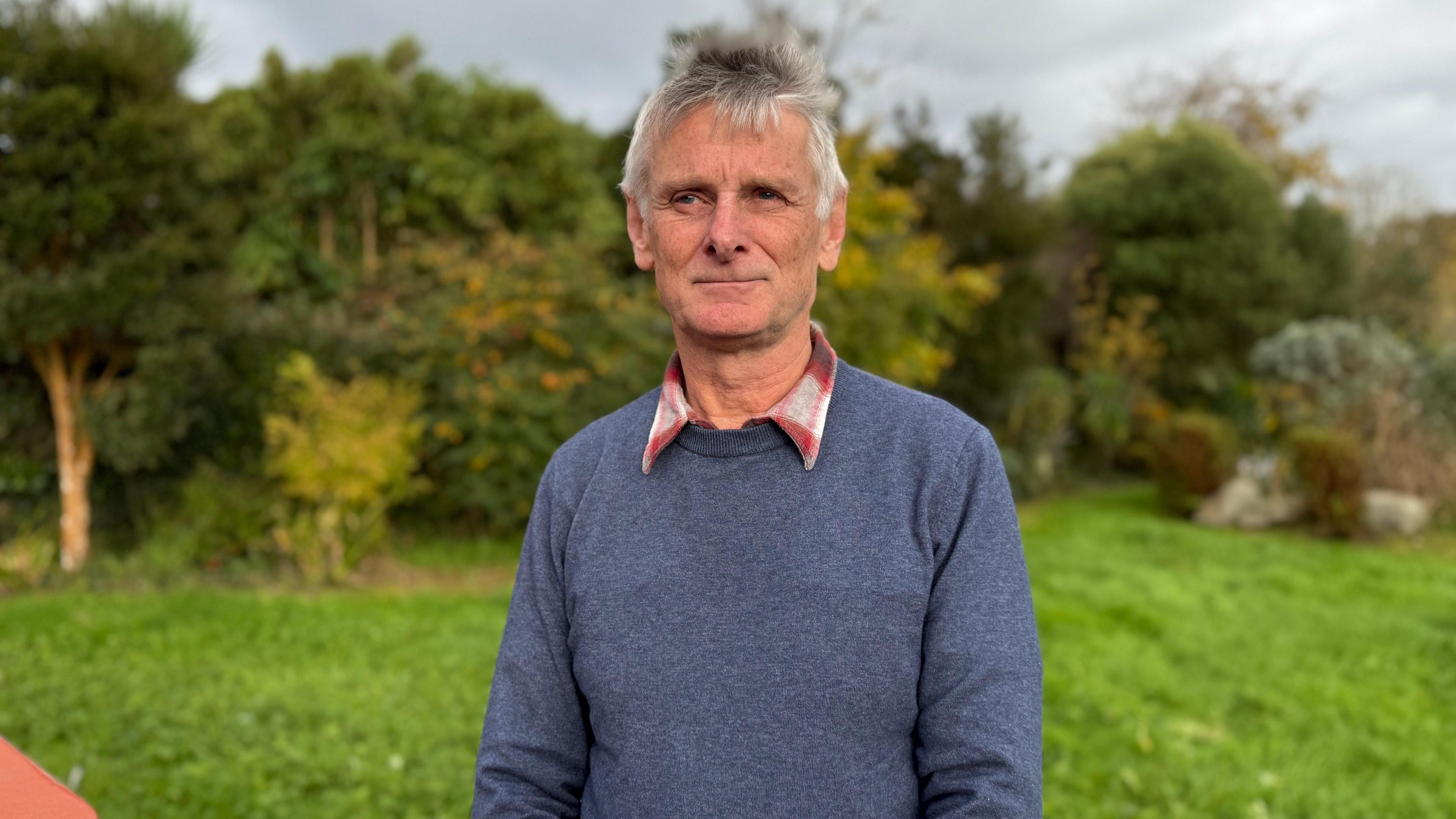 A picture of Jeff Guilbert, who is an older man with short grey hair. He is wearing a blue sweater with the collar of his read and white shirt showing. The background shows grass, trees and a grey sky.