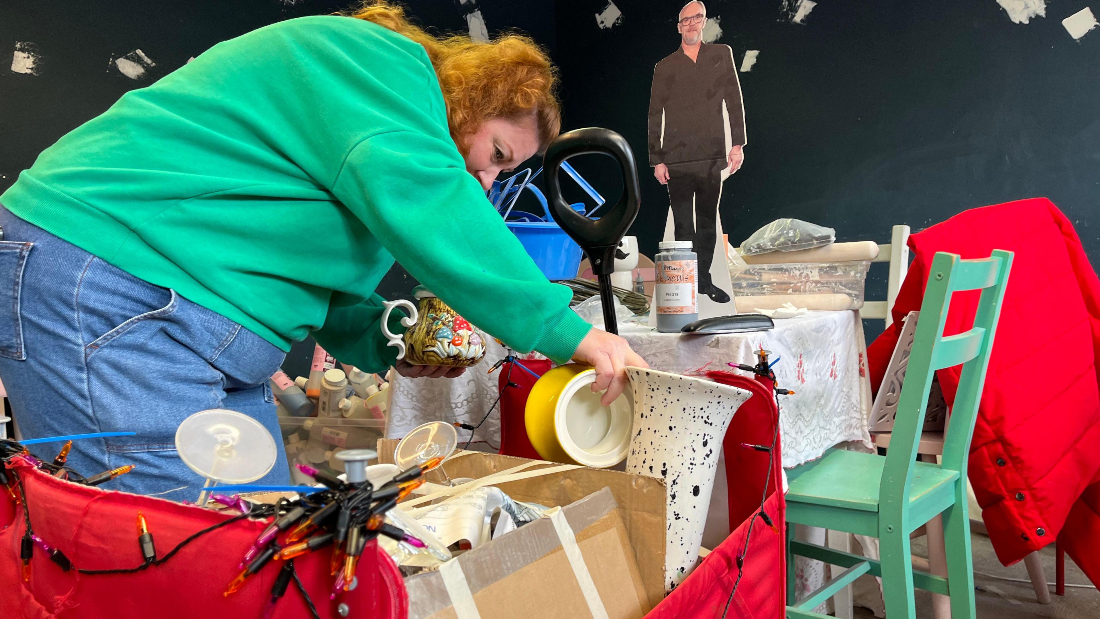 Danielle, who has ginger hair and is wearing a green jumper and blue jeans, is putting ceramic pots into a four-wheeled trolley which is covered in red cloth and fairy lights. Behind the trolley is a table with a white table cloth and a green wooden chair.