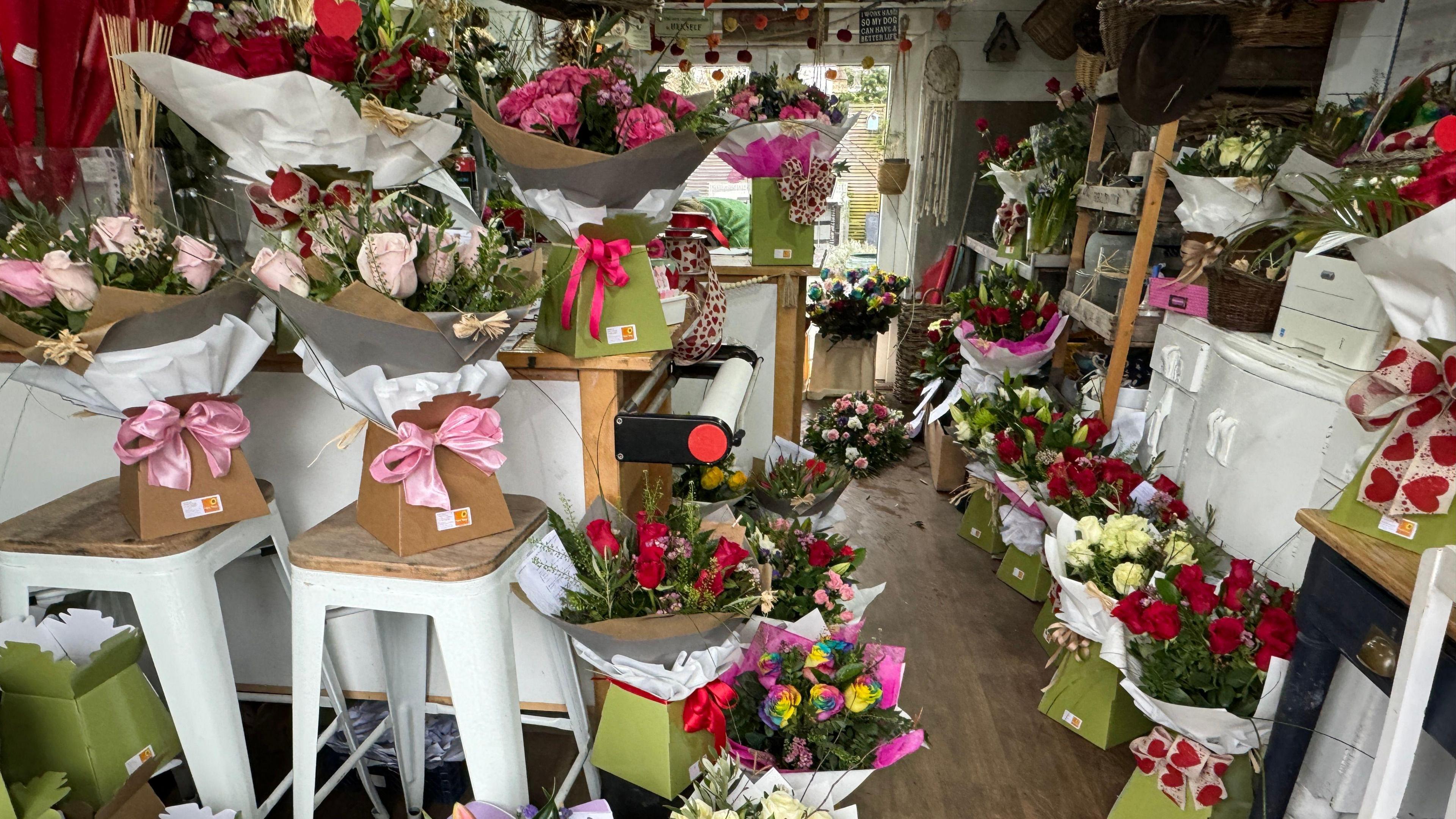 A studio with boxes and bouquets of different types of flowers like red roses with pink and red ribbons wrapped round them. 