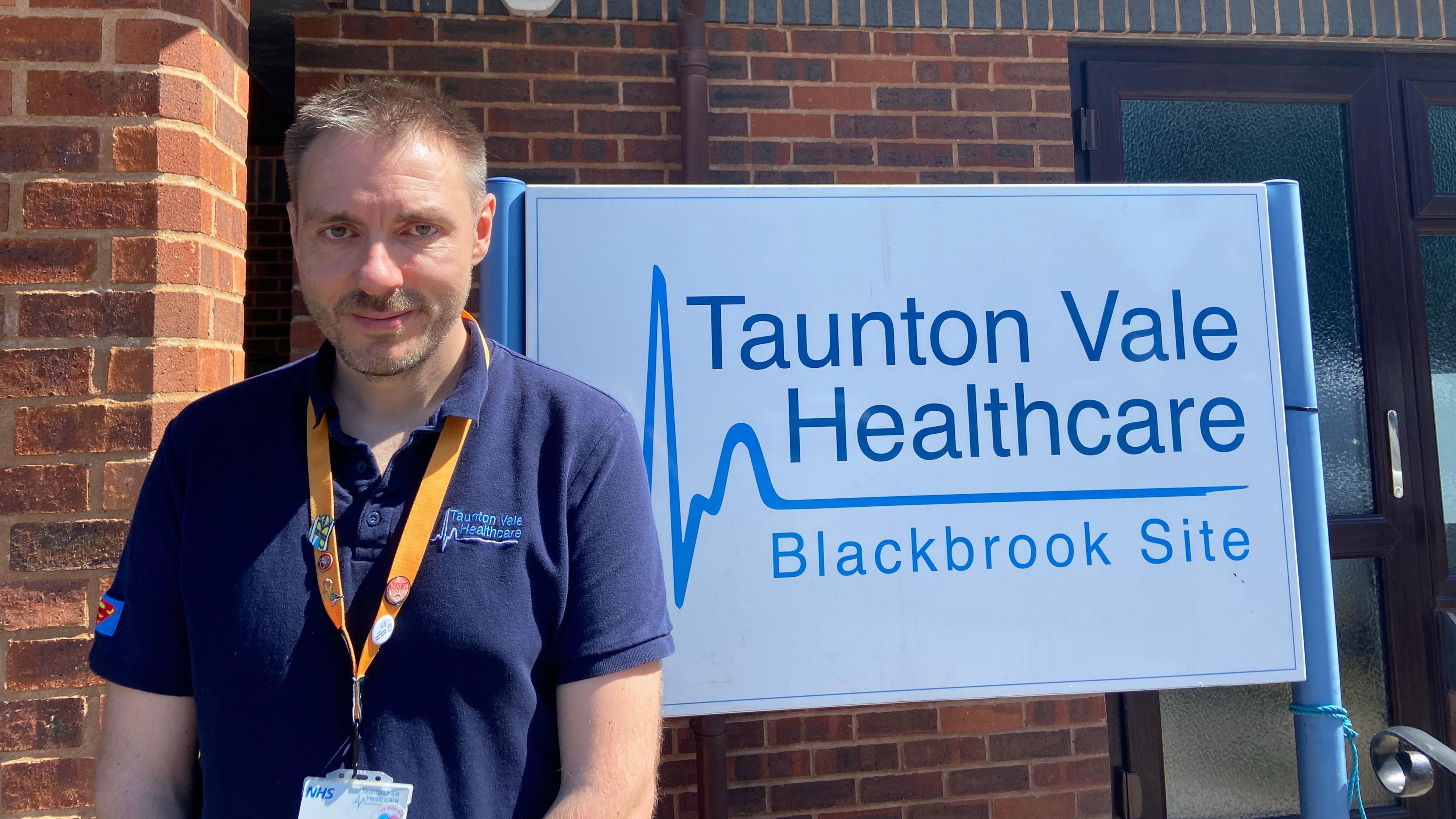 Dr Tim Norbury. He has grey short hair and is wearing a blue collared shirt underneath a khaki green jumper. He is pictured looking directly at the camera and smiling. 