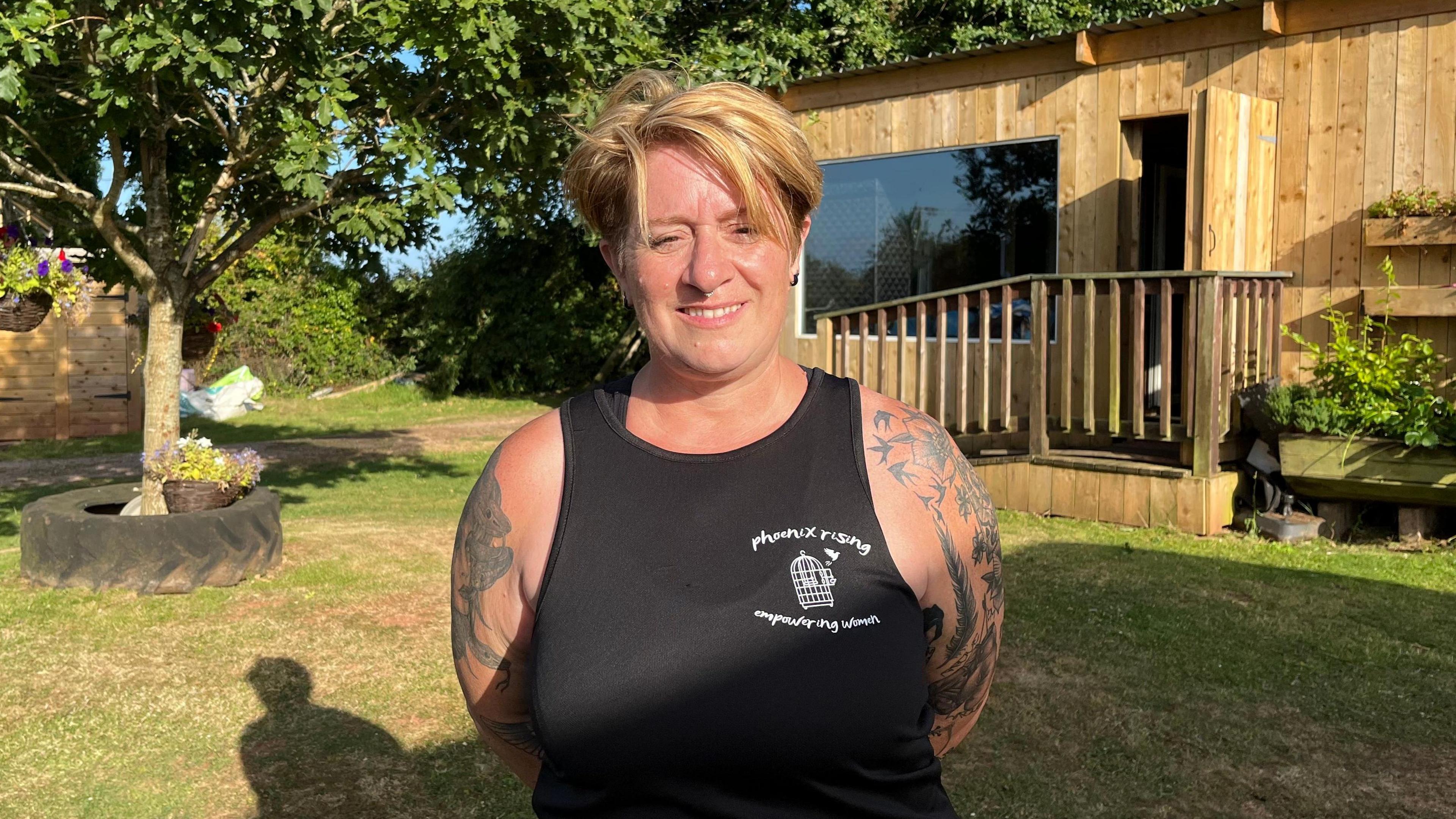 A woman with short blonde hair and a black singlet. She has tattoos on her arms. 