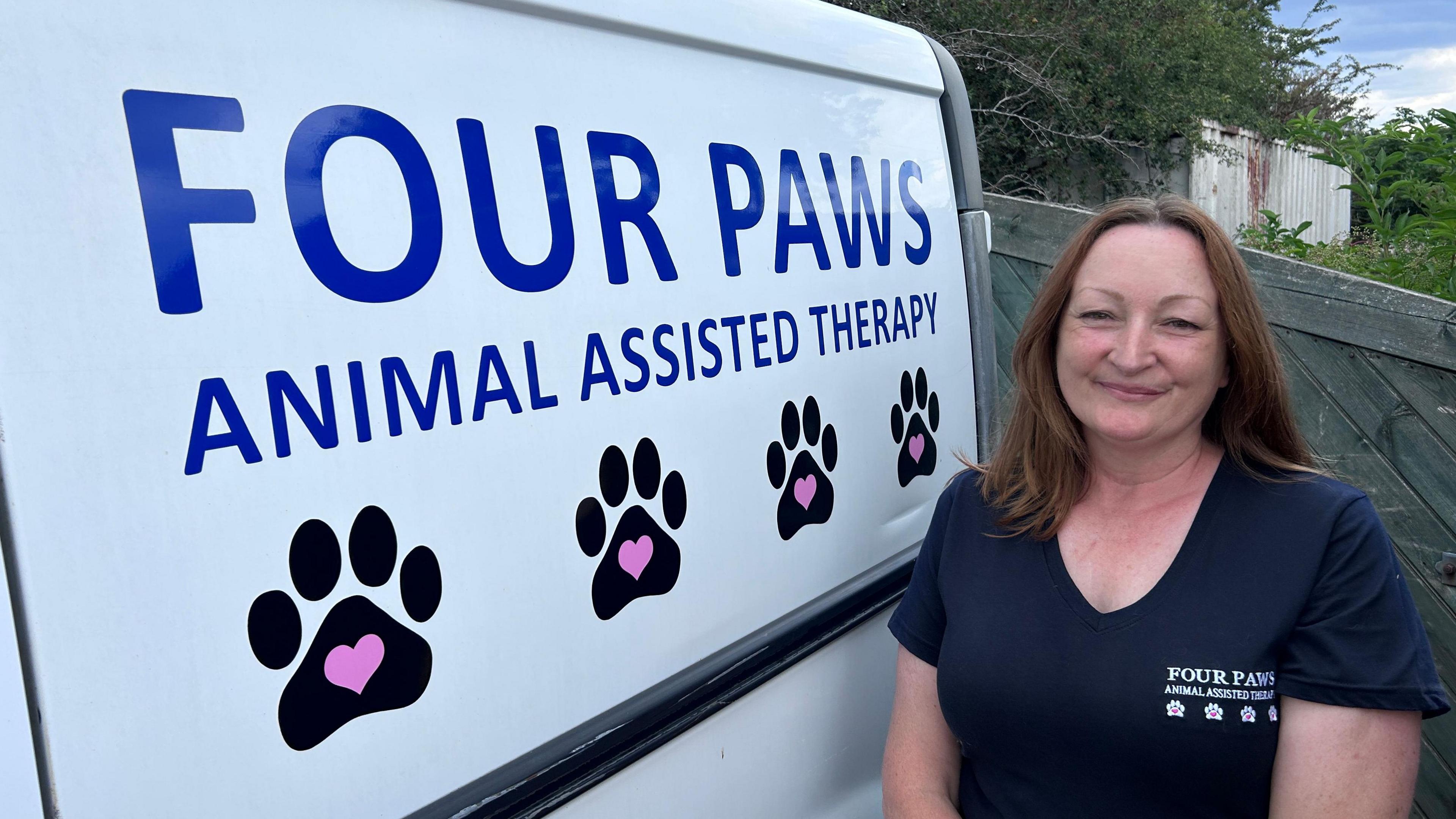 White woman with brown hair wearing a navy blue t-shirt smiling at the camera next to a white van with 'Four Paws Animal Assisted Therapy'  written on the side. 