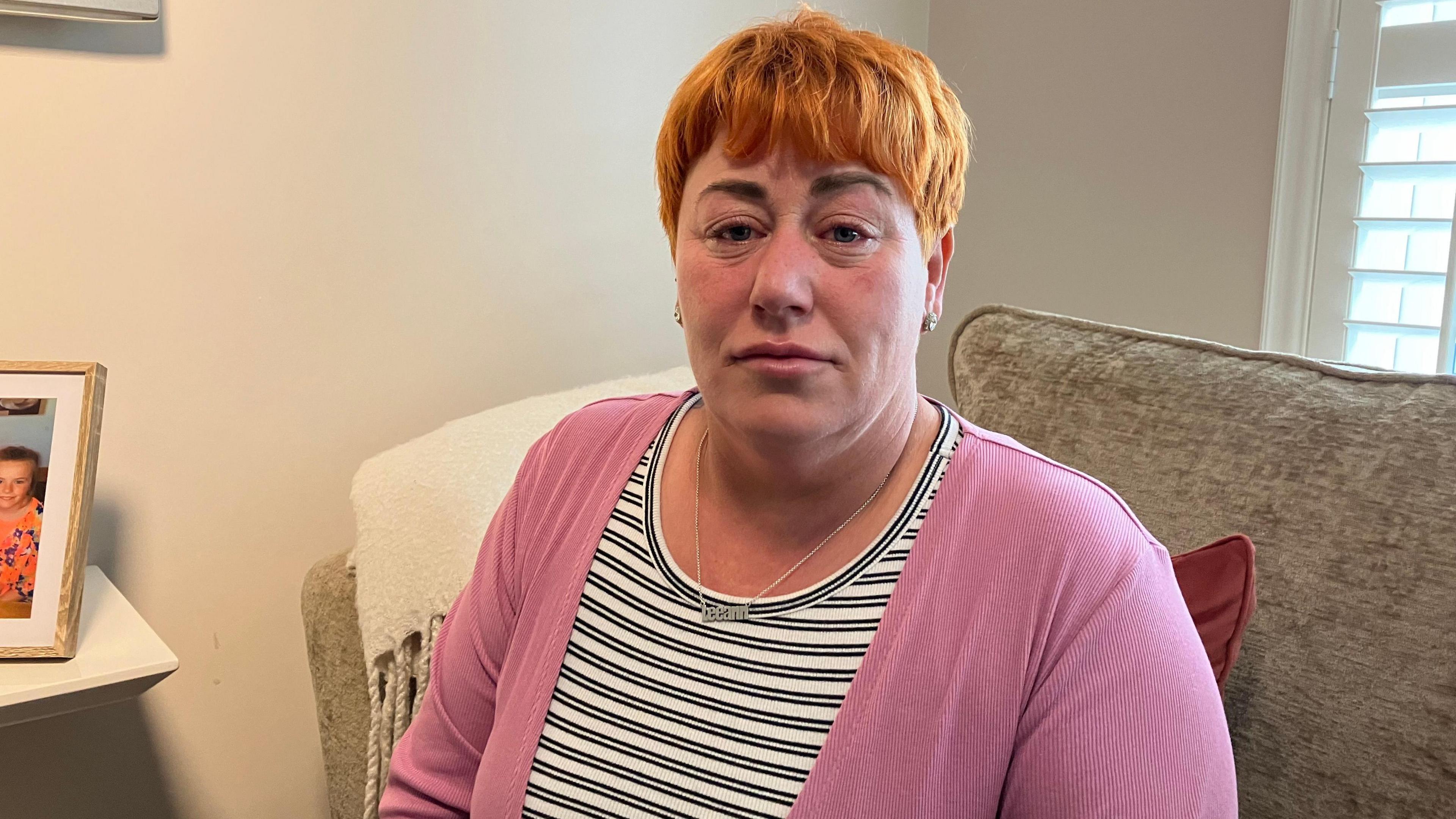 Leanne mclaughlin sits on a chair, with a picture of her family to her left and a window behind her