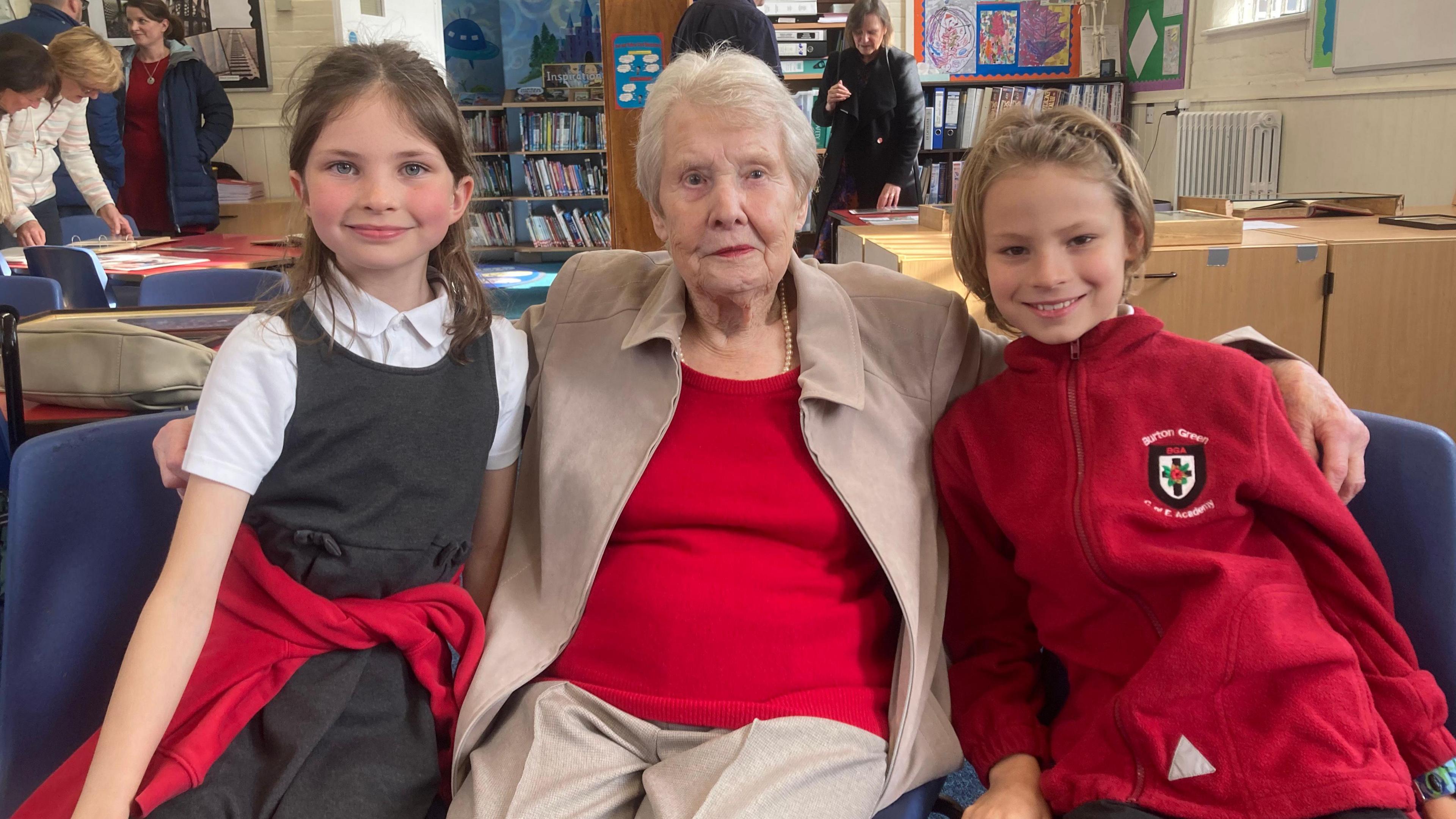 An elderly woman sits between two children. The woman has short grey hair and is wearing a red top and a beige jacket. The children are wearing red and grey school uniforms.