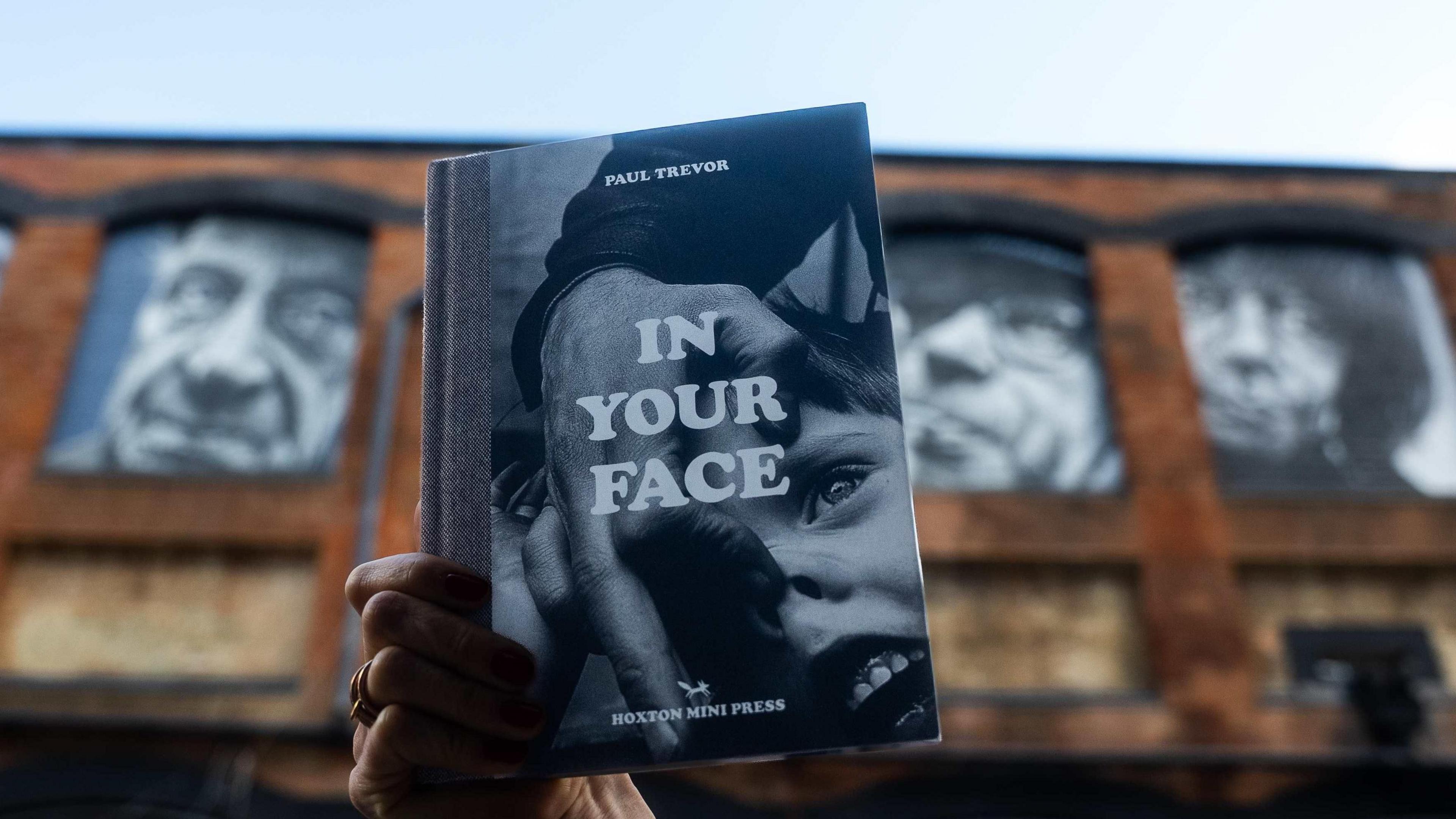 At the front of the image, we can see Paul Trevor's book In Your Face and in the background, three images from the mural are blurred.