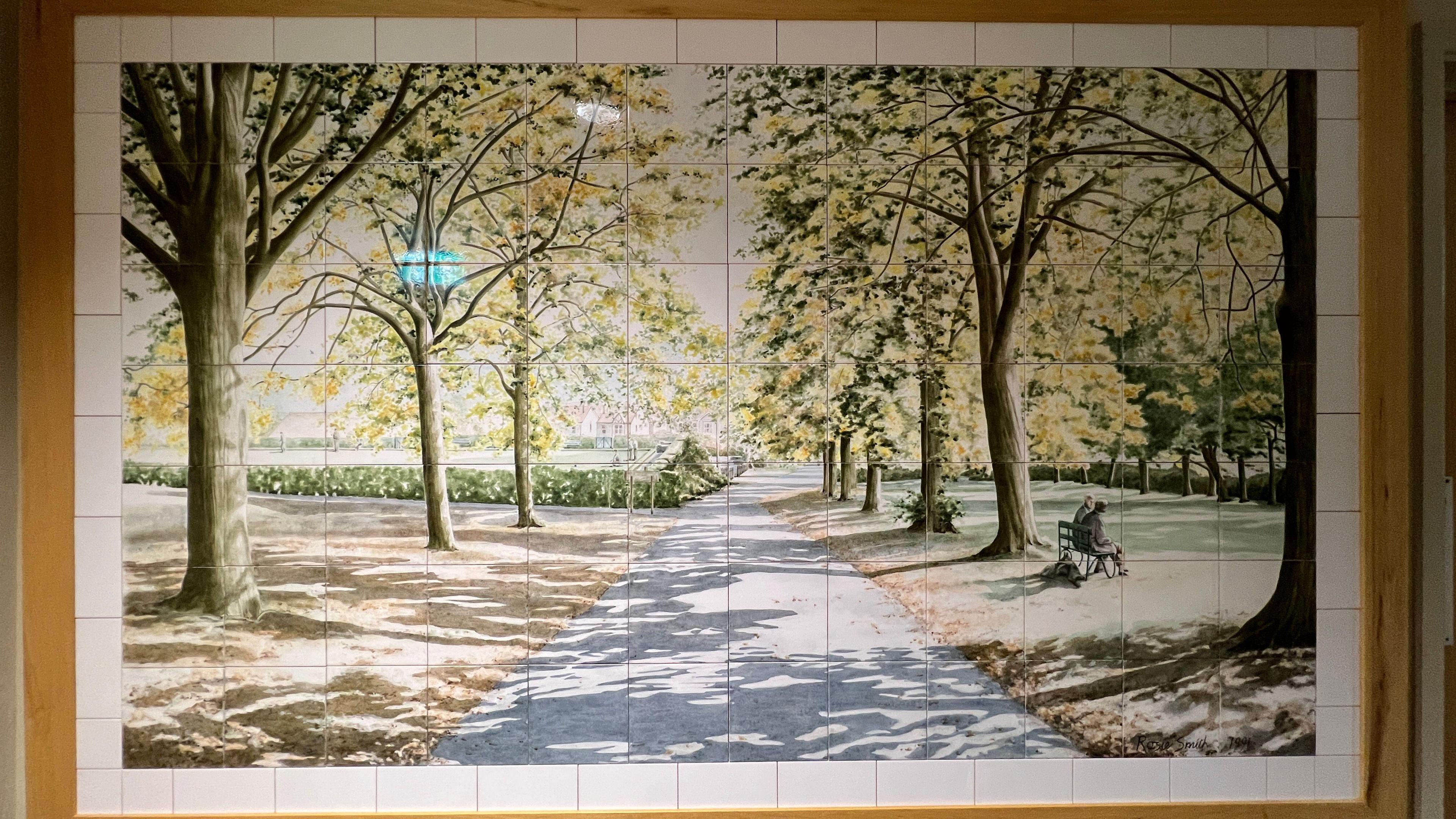 Framed painting on tiles of a park with trees and a footpath, with sun shining through the leaves and onto the ground below. A couple sits on a bench to the right, enjoying the view