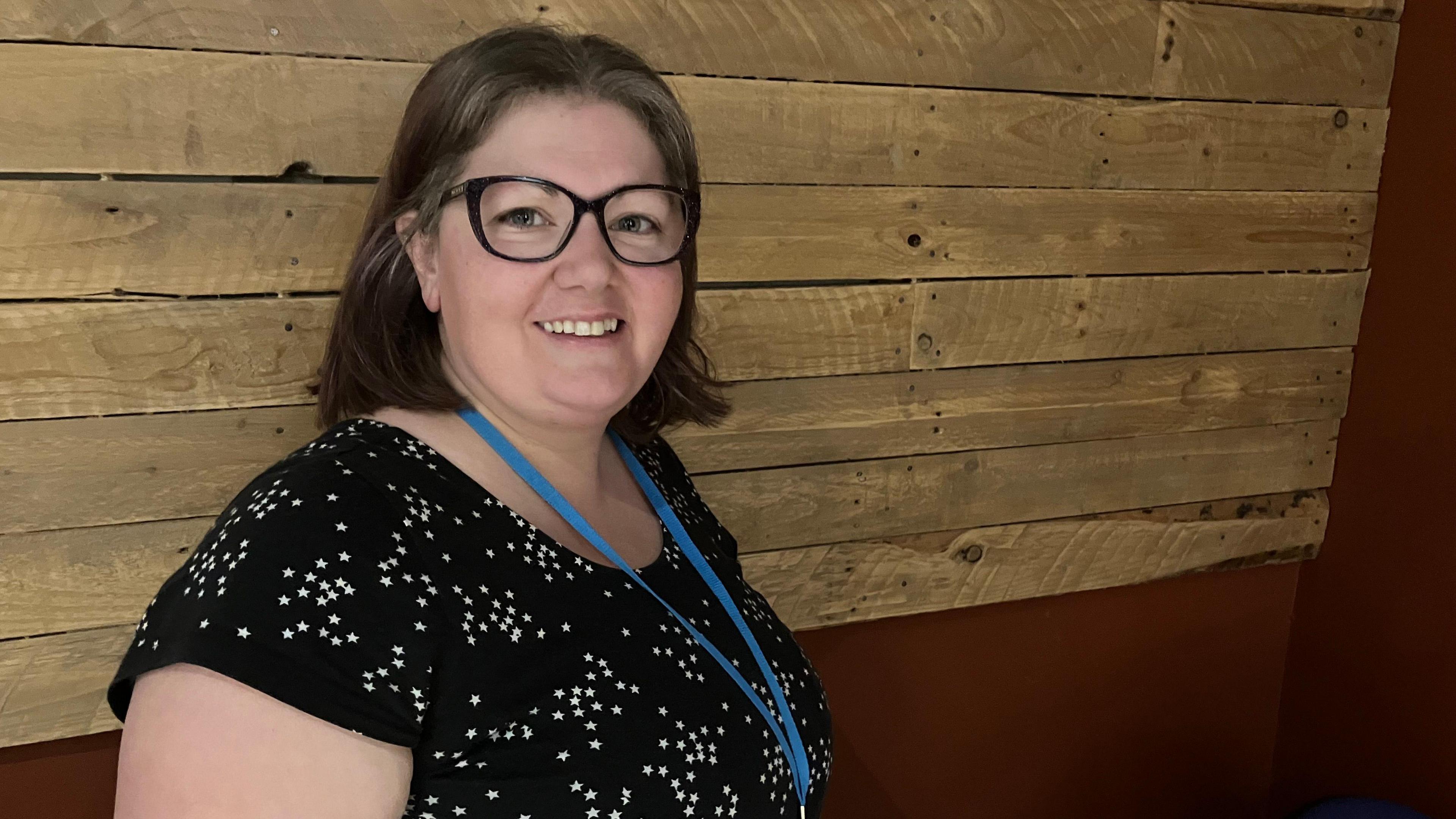 A smiling Cat Hemmington looks directly at the camera from the side, she is wearing a black dress with white stars on it. She has a blue lanyard and black glasses. 