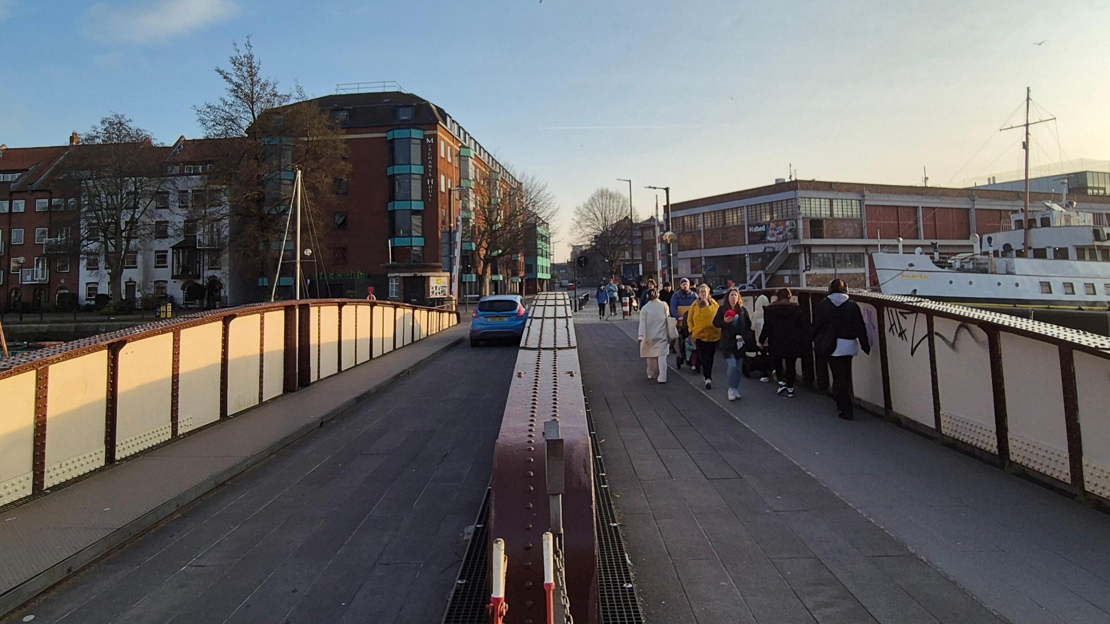 A view across the Prince Street Bridge. On the left a blue car is driving across the bridge, on the right people are walking. There is a ship and buildings in the background.