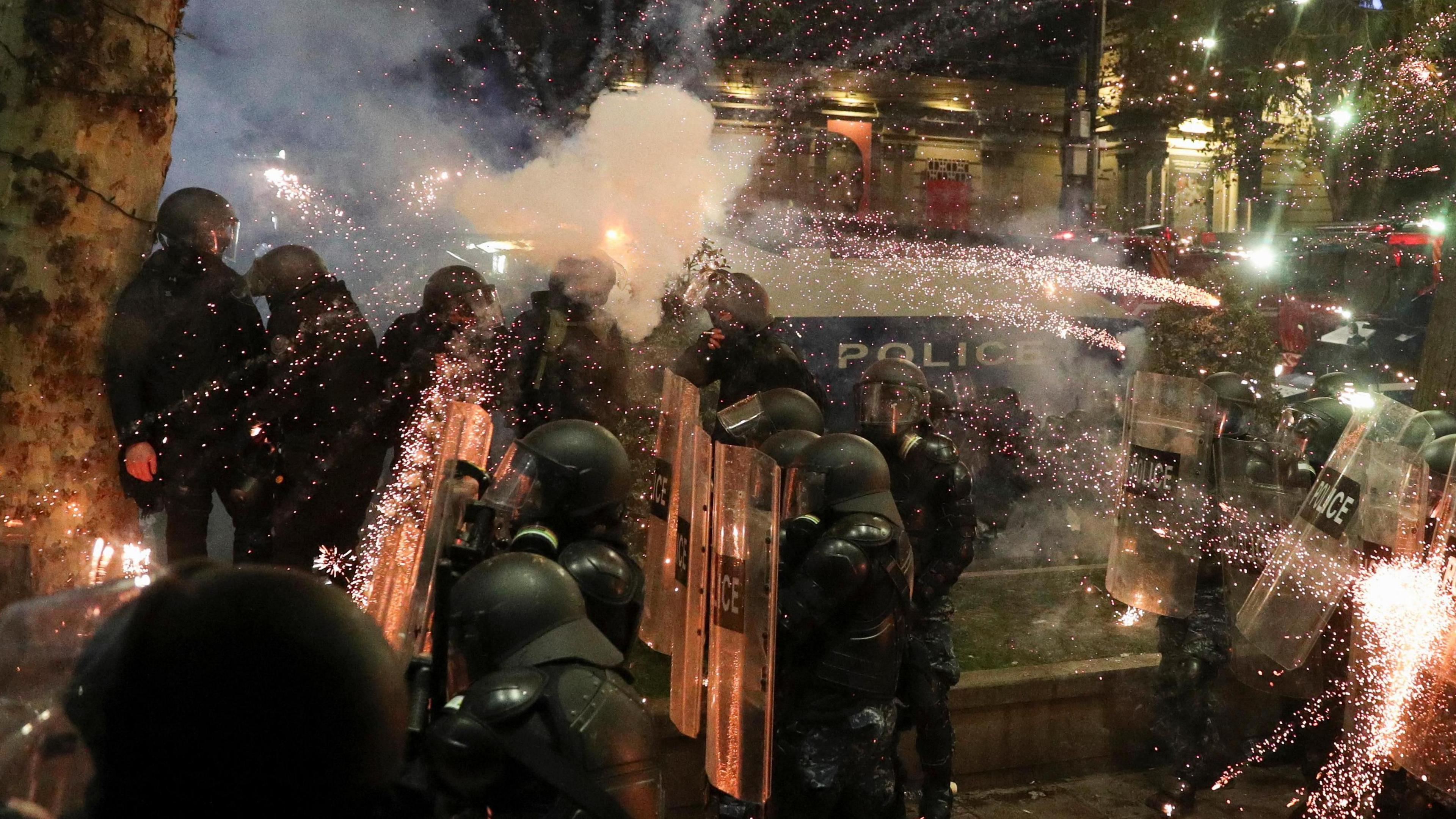 A firework explodes in a shower of yellow and red sparks above the heads of a group of riot police officers. They are wearing black uniforms and heavy riot helmets, their faces obscured. Several are holding clear plastic shields.