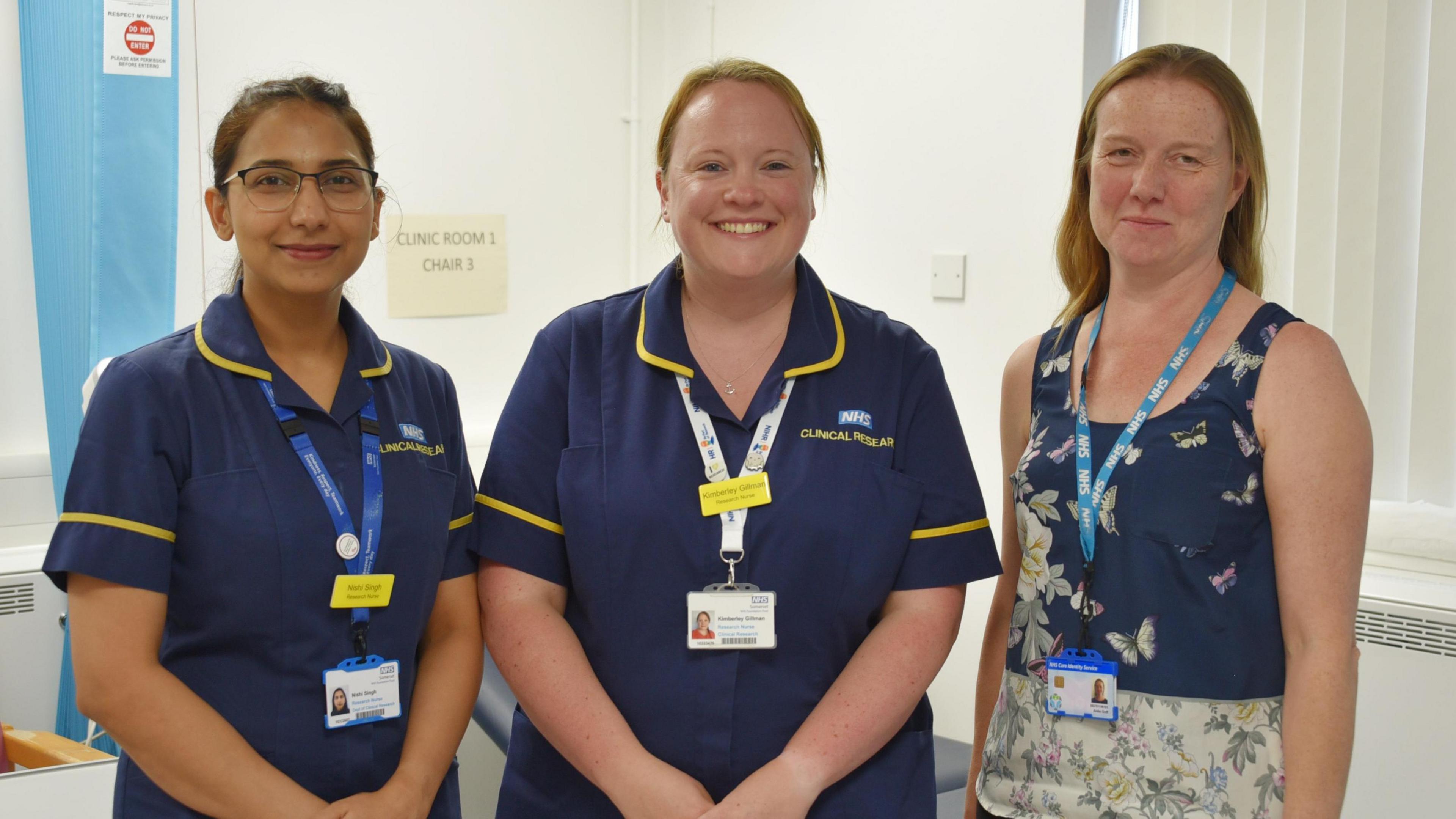 left to right - Nishi Singh and Kimberley Gillman and Dr Anita Goff. Nishi and Kimberley are both wearing nurse's overalls, whike Anita has a flowery top on. They are standing in a hospital setting with a blue medical curtain behind Nishi