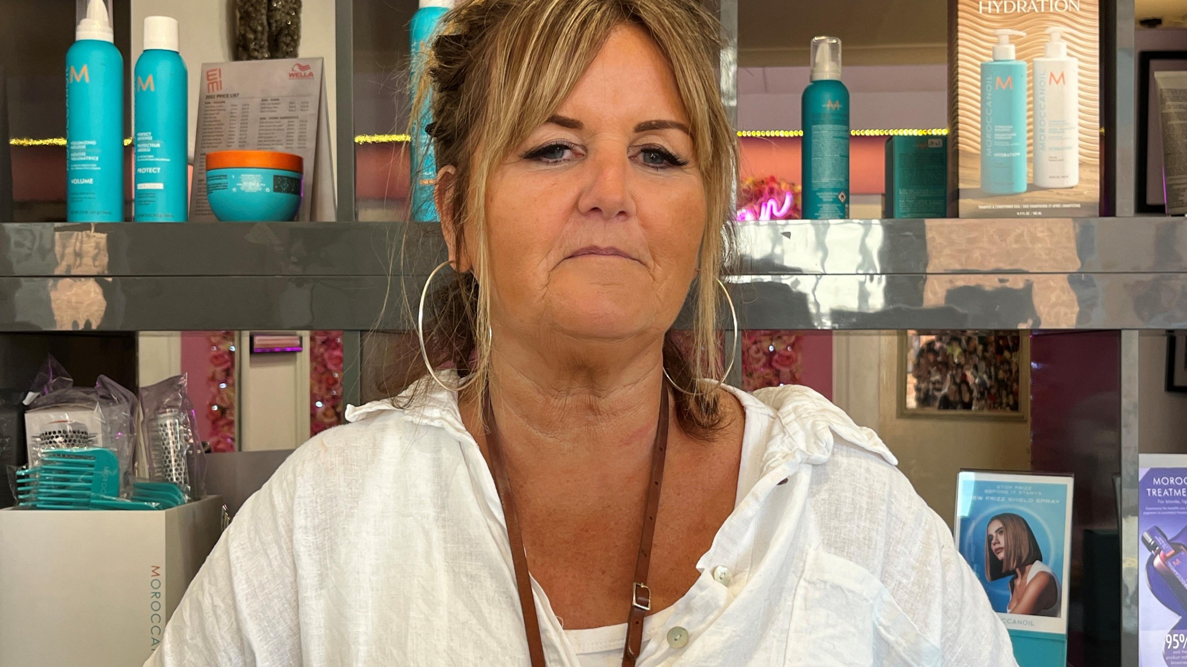 Hairdresser Debbie Lee is wearing a white blouse and is standing in front of a shelf full of hair products in her salon