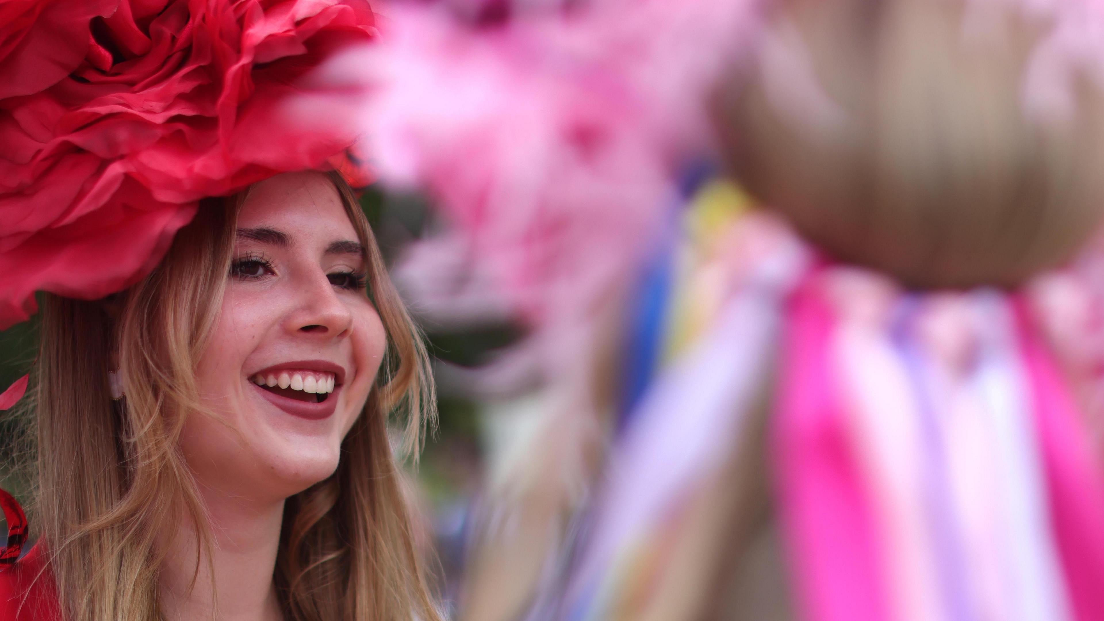 A young woman wearing a red hat smiles with another woman in a pink headdress in soft focus in the foreground