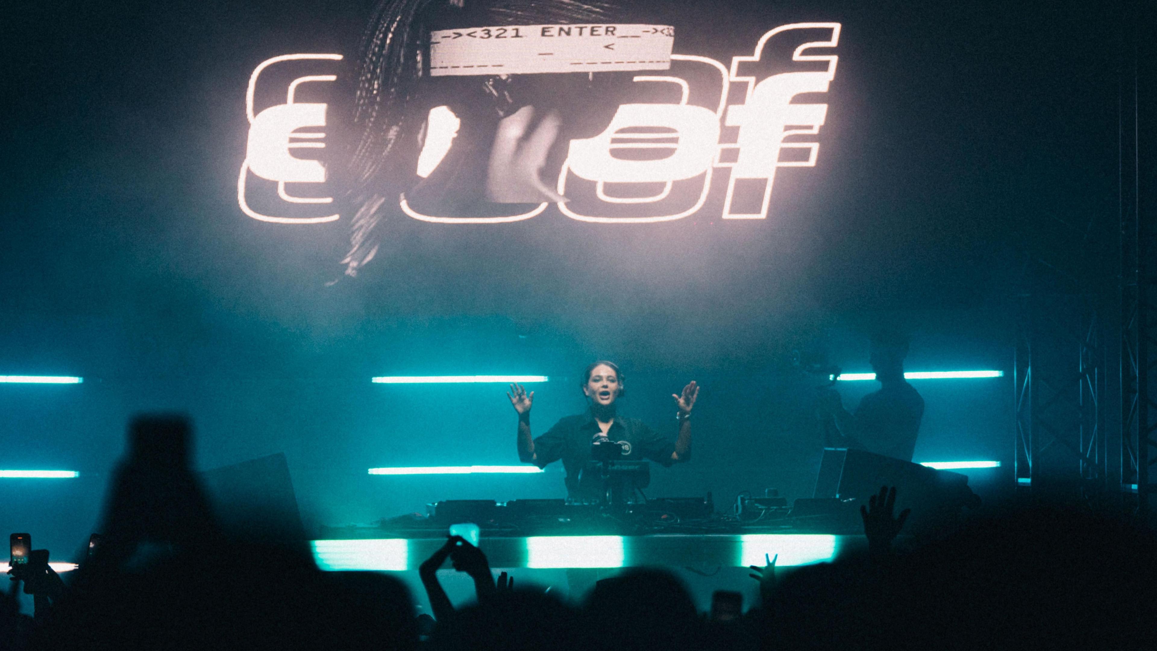 A woman DJs behind decks to a busy crowd. The word 'doof' is behind her. The stage lighting is blue and turquoise.