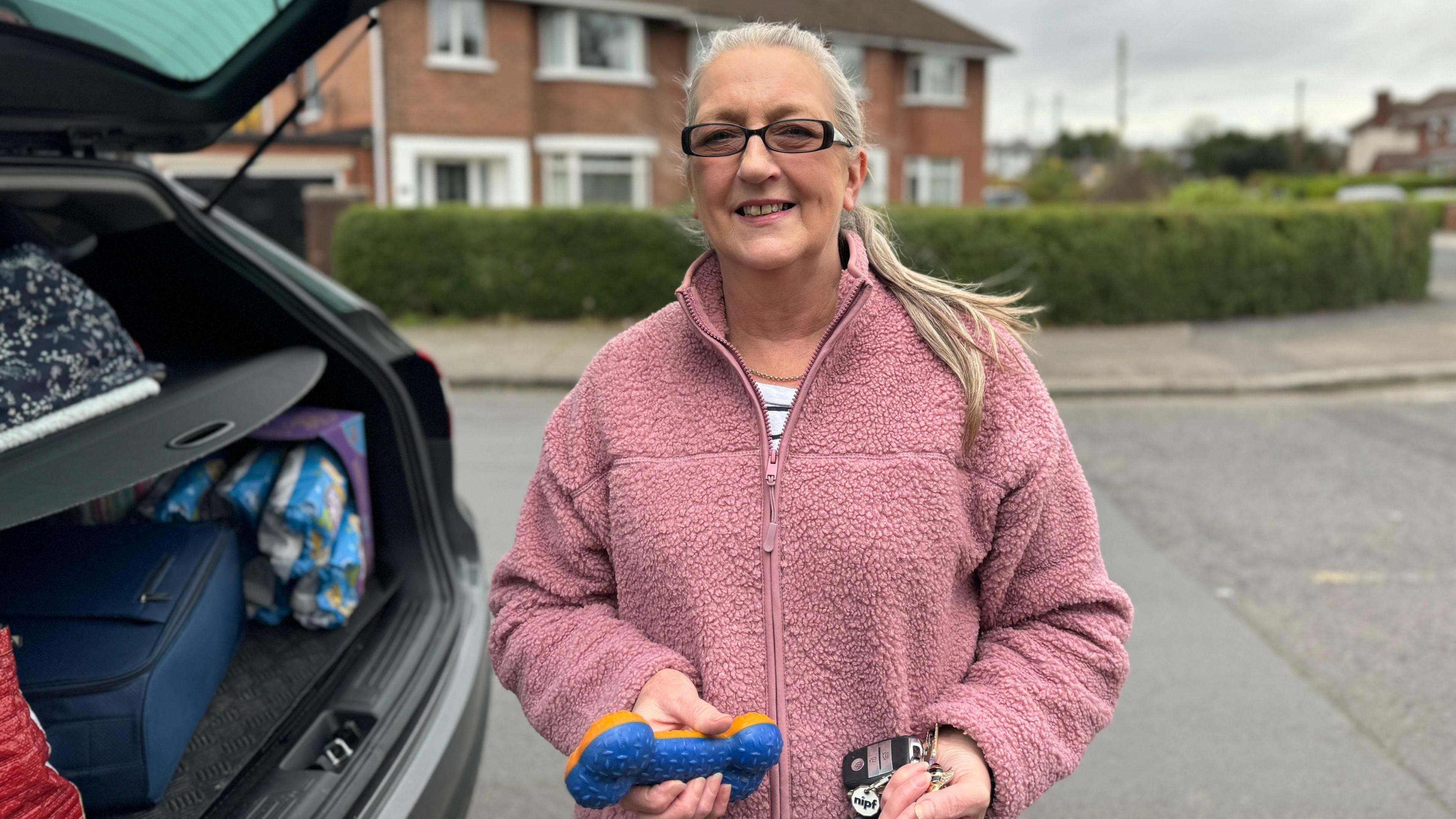 Suzie Stewart is wearing a light pink fleece and has long grey hair tied up. She is wearing dark framed glasses and is holding a blue and orange bone dog toy.